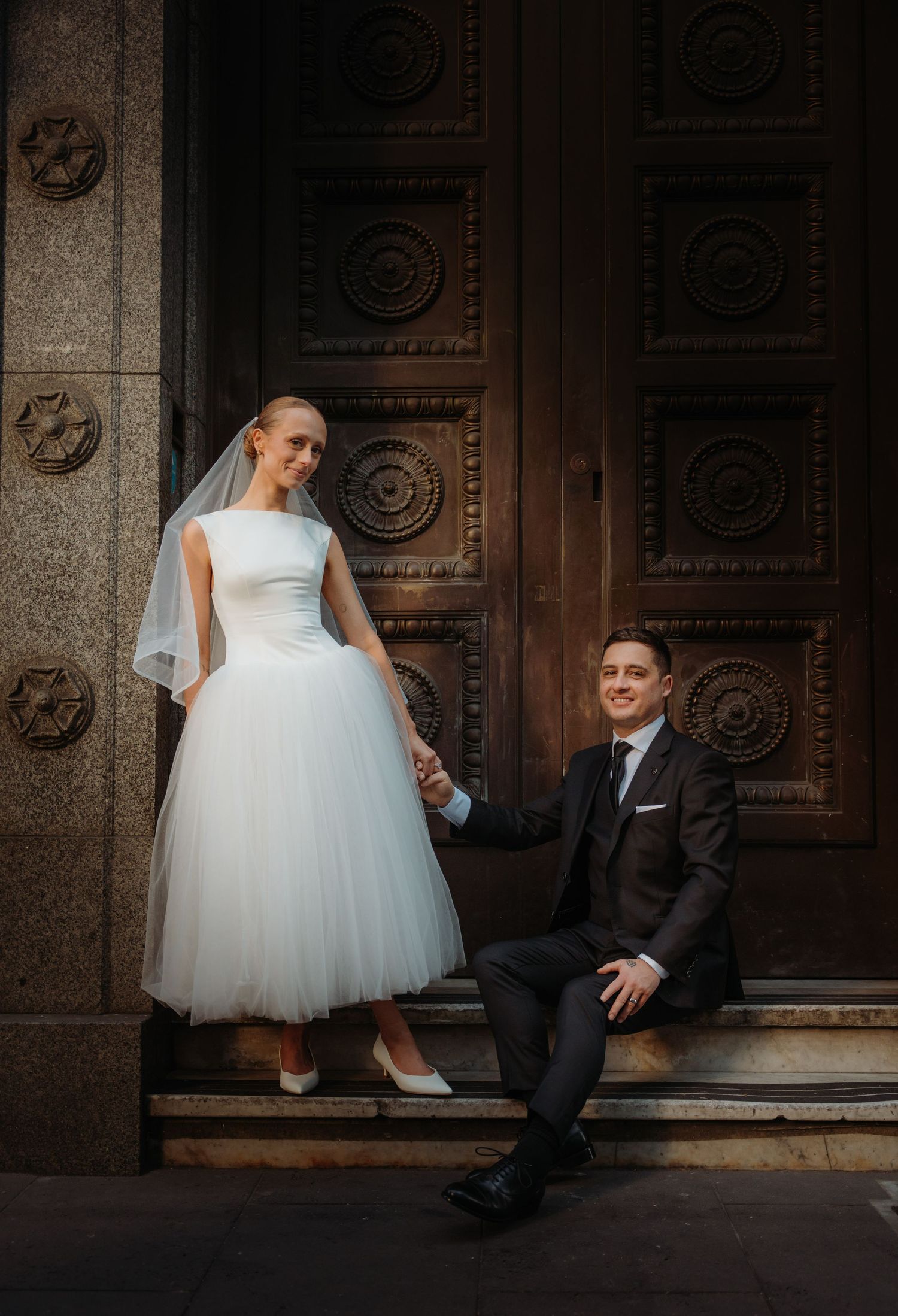 Bride in tea-length tulle gown and groom in dark suit pose on stone steps before ornate wooden doors.