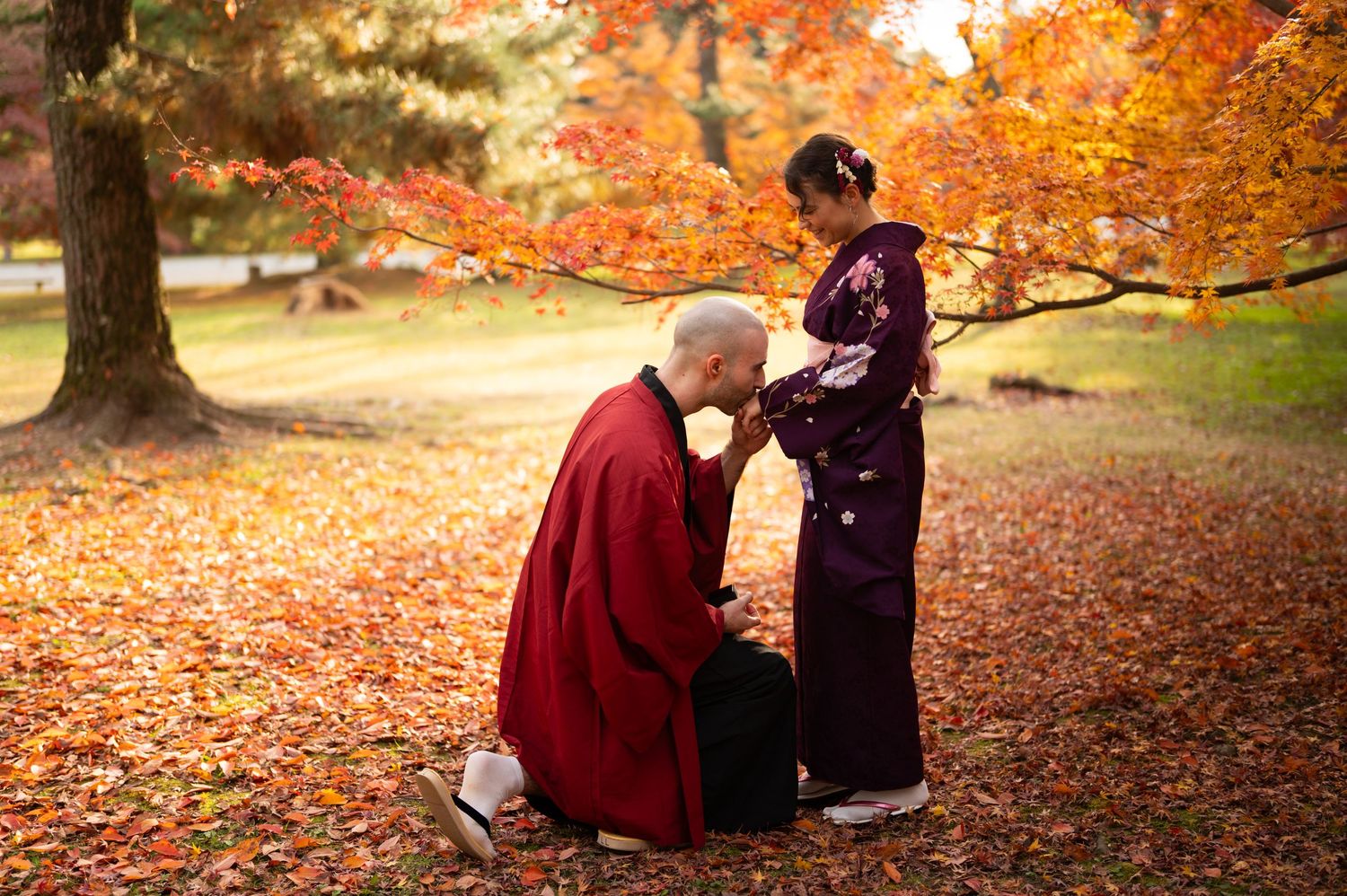 Man in red kimono kneels on autumn leaves holding hands with a woman in a purple kimono beneath vibrant orange maple trees.