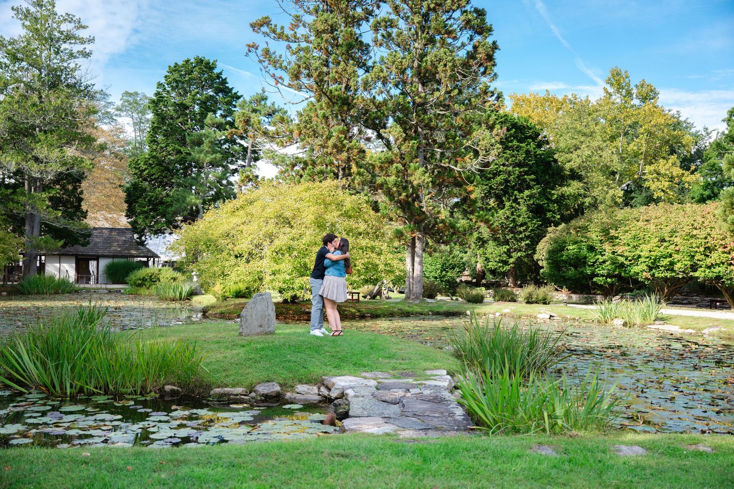 Couple embracing near a tranquil pond in a lush Japanese garden with green trees and manicured lawns in Westchester County after a proposal.
