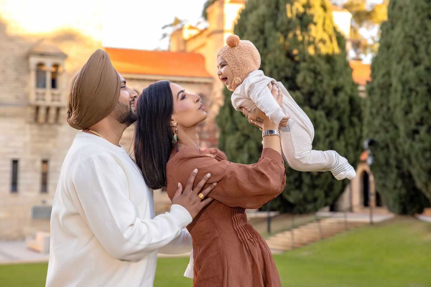 Smiling couple in traditional and modern attire lifts a baby in a bear costume outdoors near a historic stone building and tall green trees.