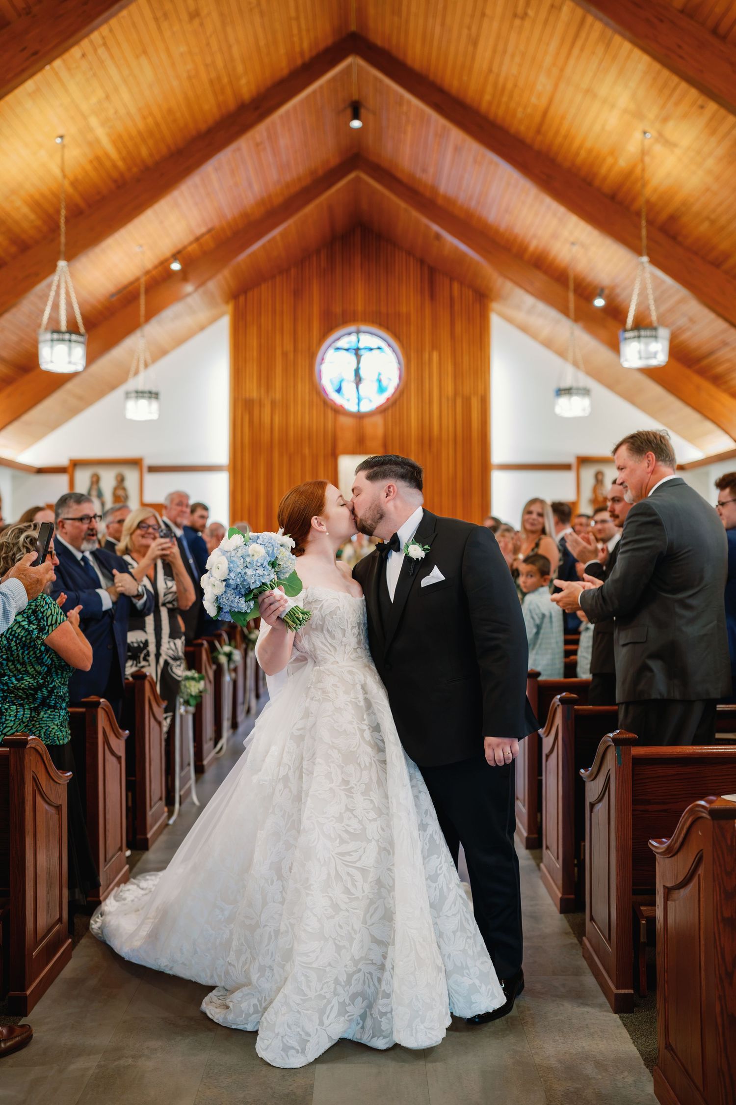Bride and groom share first kiss as newlyweds in a charming wooden chapel aisle, surrounded by cheering guests.