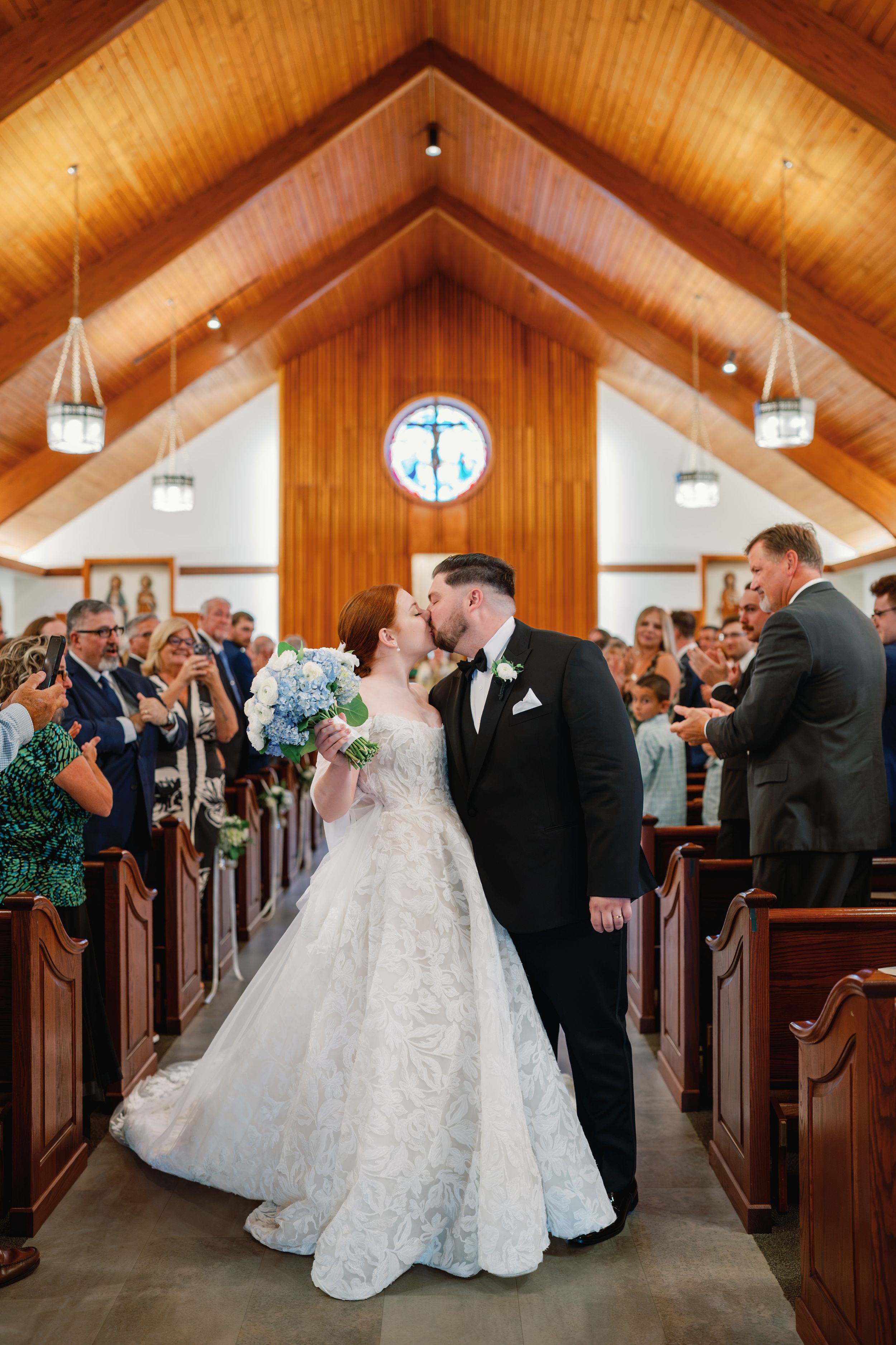 Bride and groom share first kiss as newlyweds in a charming wooden chapel aisle, surrounded by cheering guests.