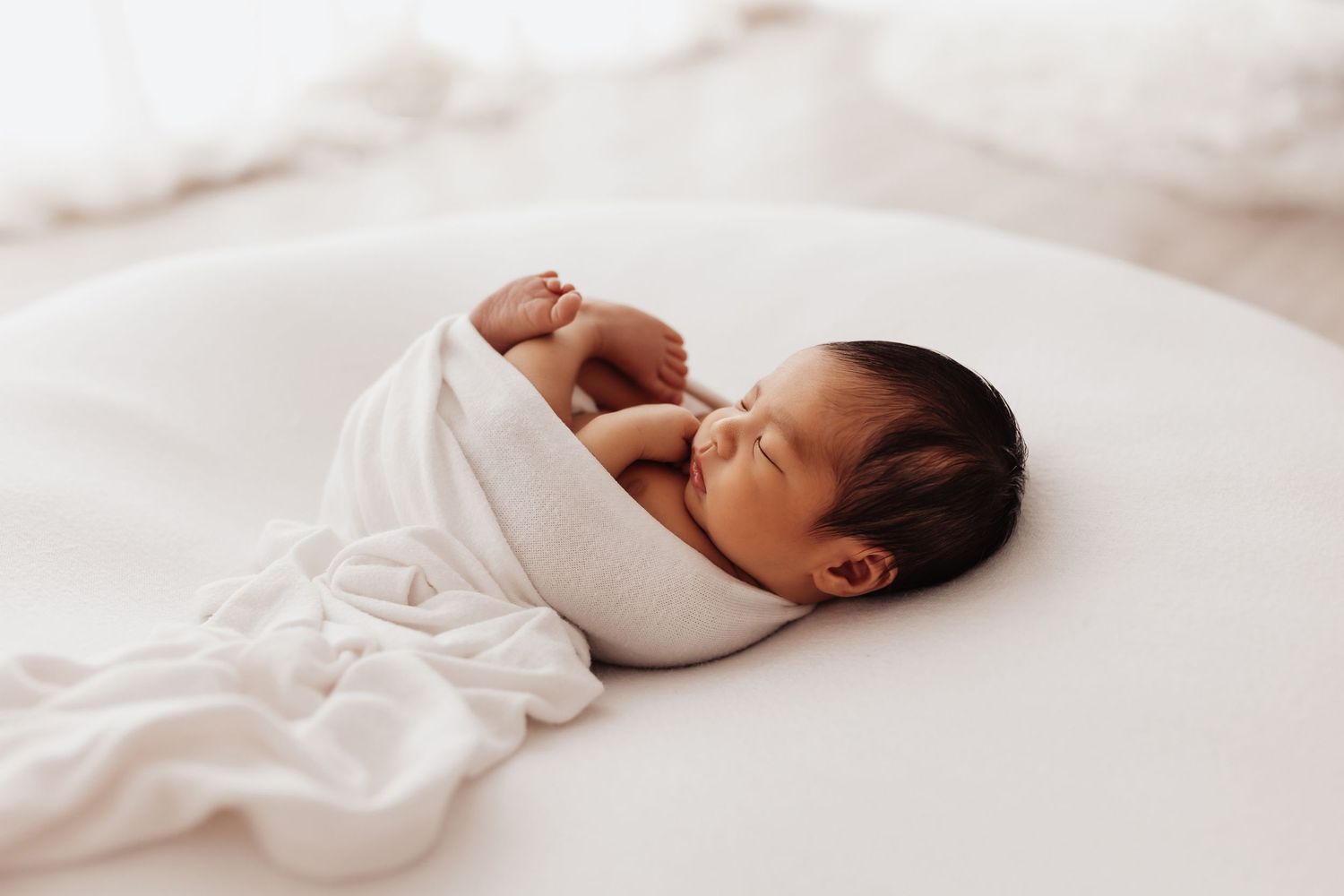 Newborn baby wrapped in soft white swaddle blanket sleeping peacefully on light colored bedding.
