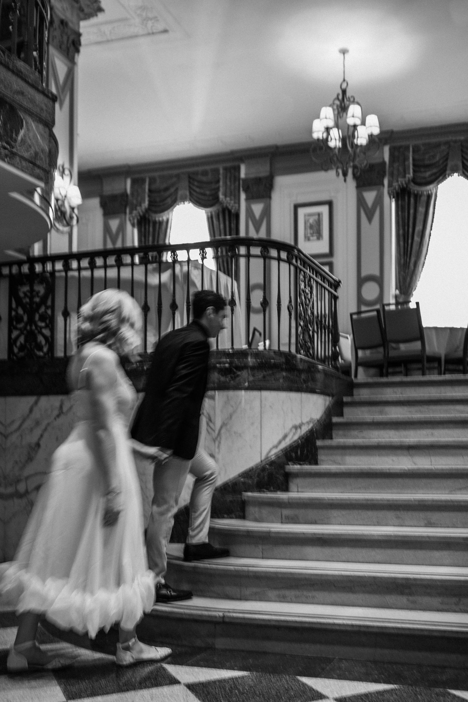 A black and white photo captures a couple ascending elegant stairs in a vintage-style interior with chandeliers.