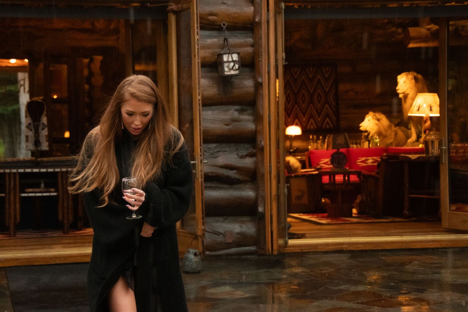 A person in a black coat stands outside a cozy log cabin restaurant with warm lighting visible through windows at dusk.