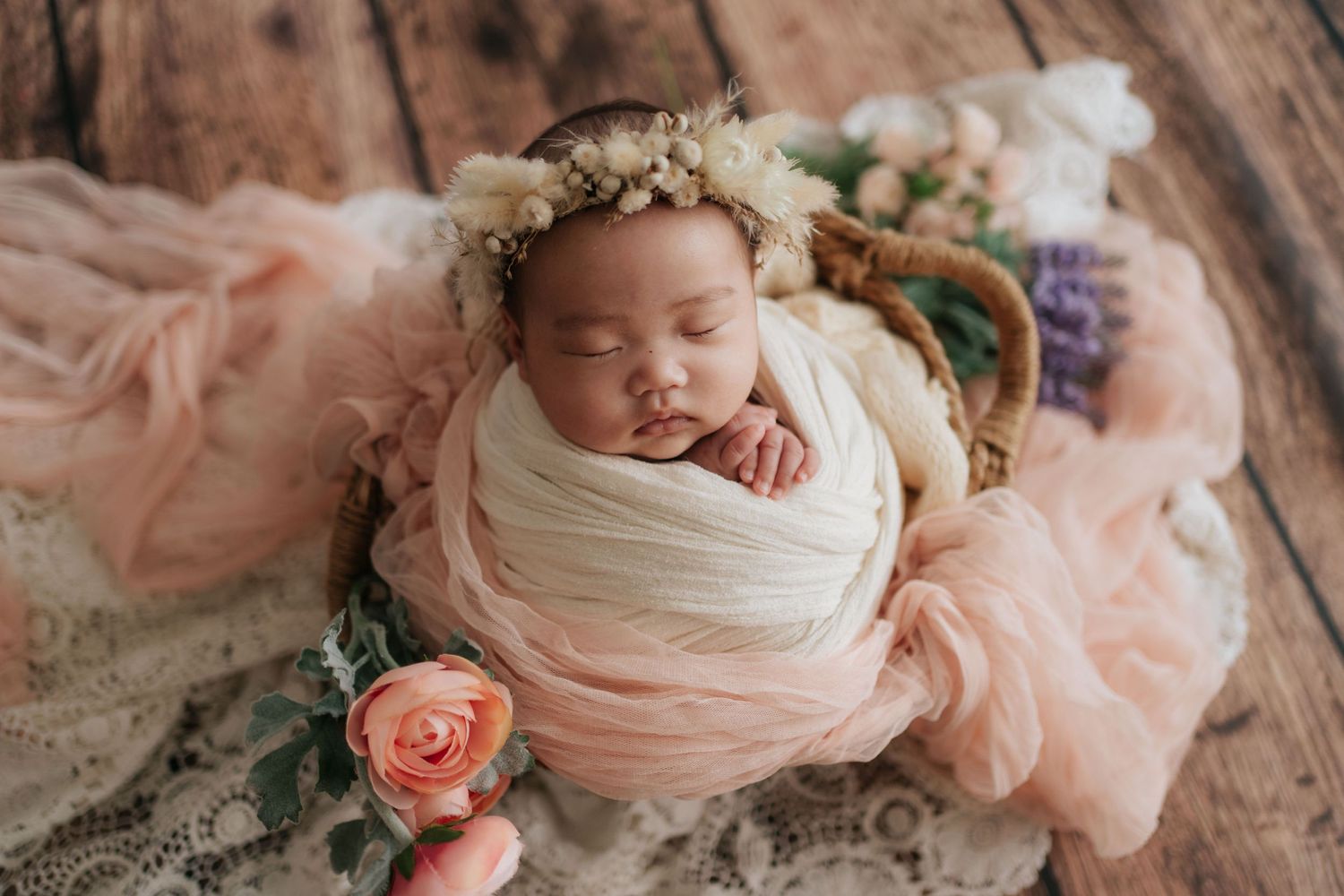 Sleeping newborn wrapped in pink fabric with floral crown and roses on wooden surface.