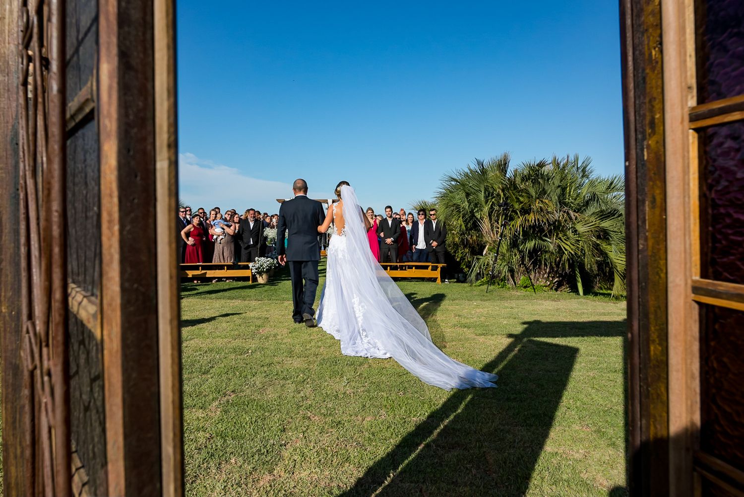A wedding ceremony takes place outdoors with a flowing white veil and palm trees in the background.