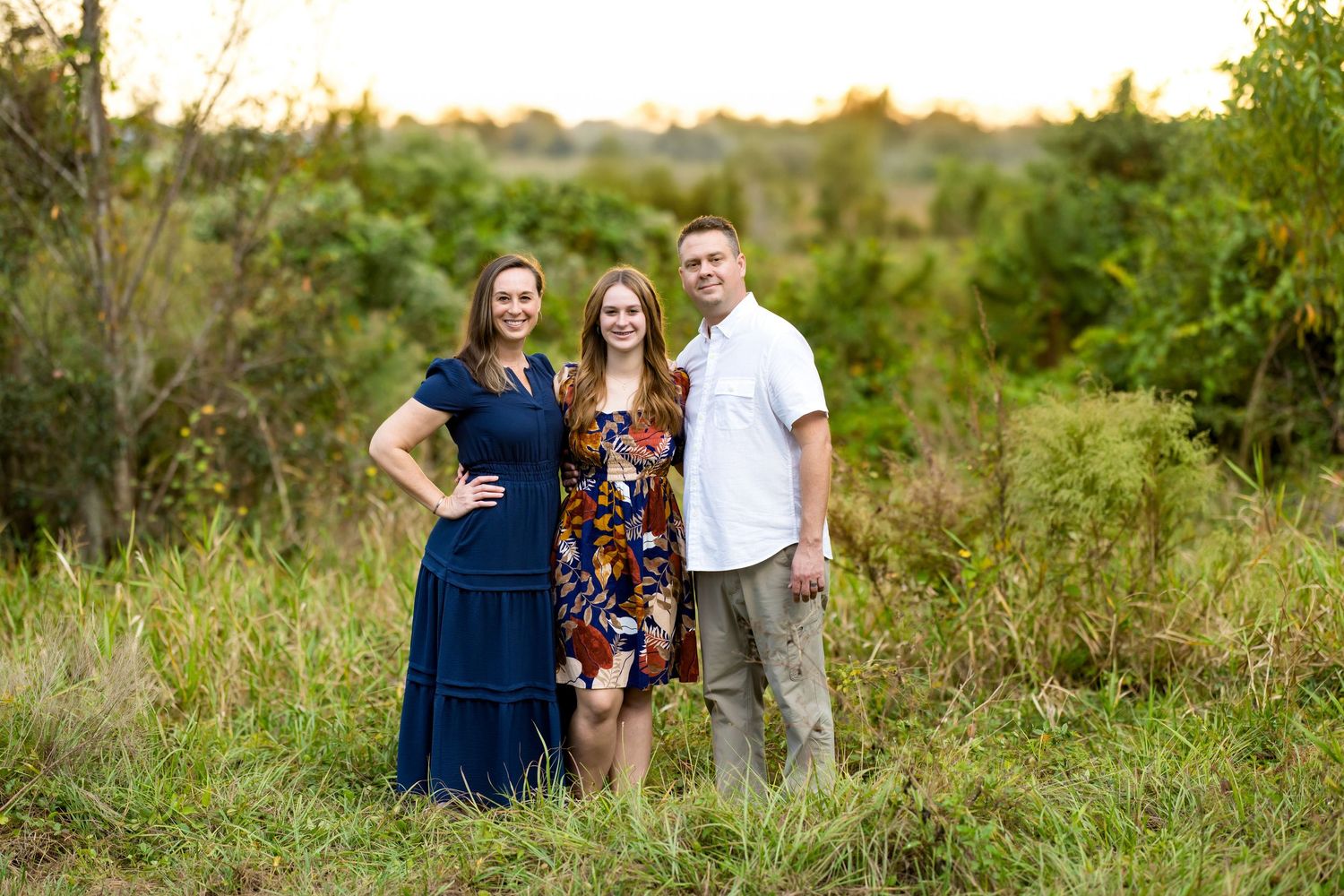 Three people stand together smiling in a grassy field during golden hour.