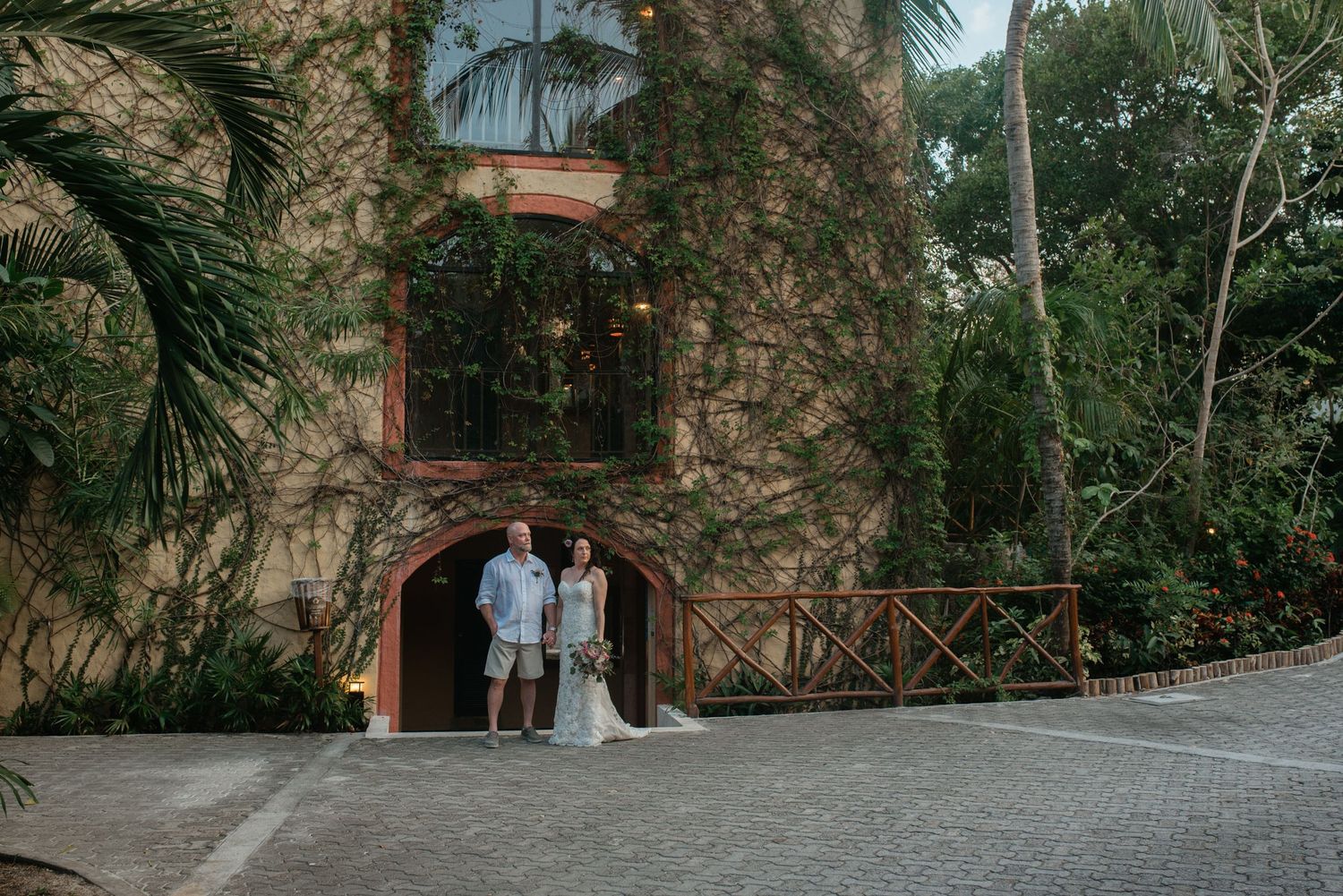 A couple in wedding attire stand together between rustic stone walls in a scenic garden pathway.