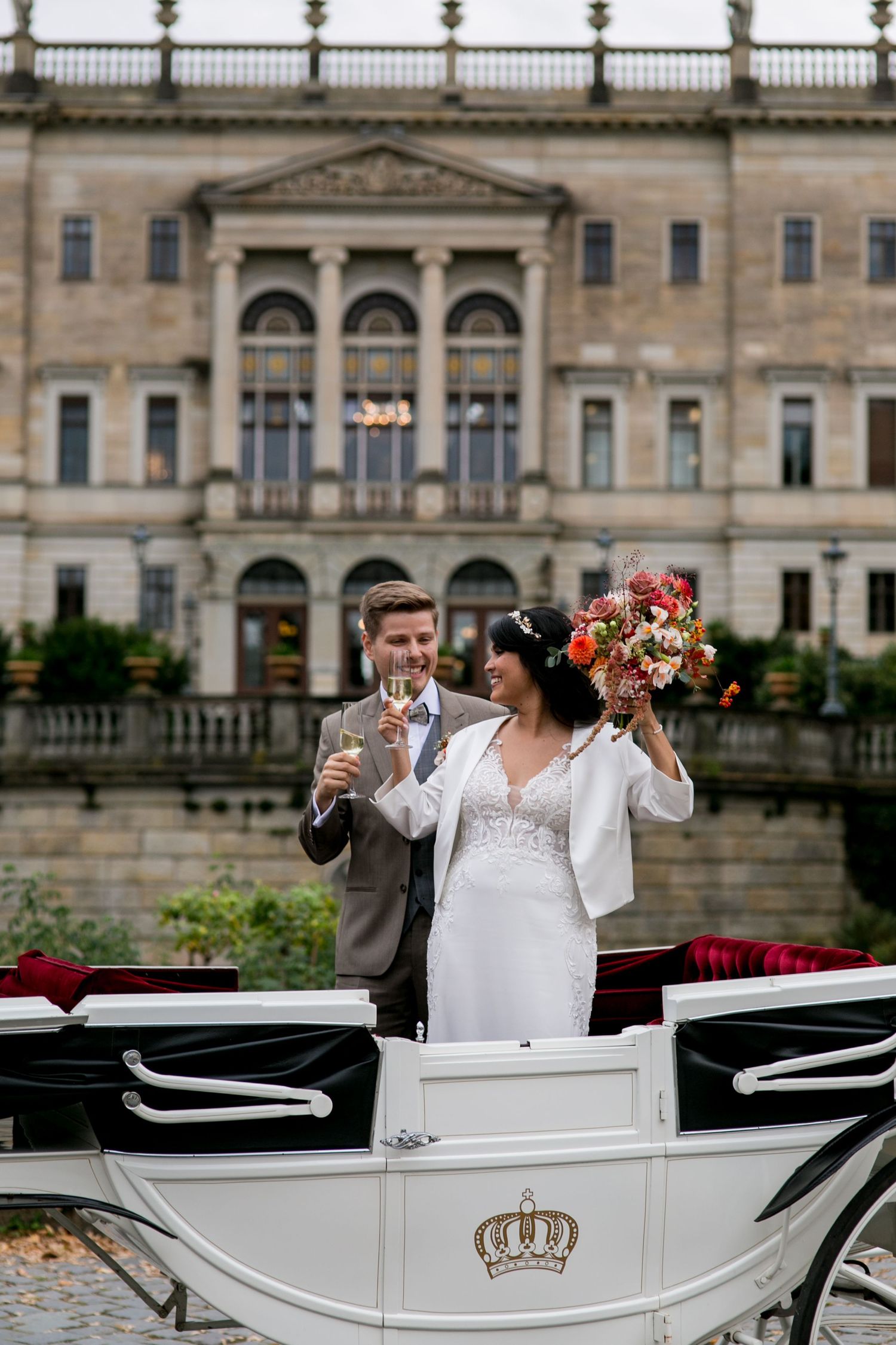 A wedding couple celebrates with champagne beside an elegant white horse-drawn carriage in front of a historic mansion.