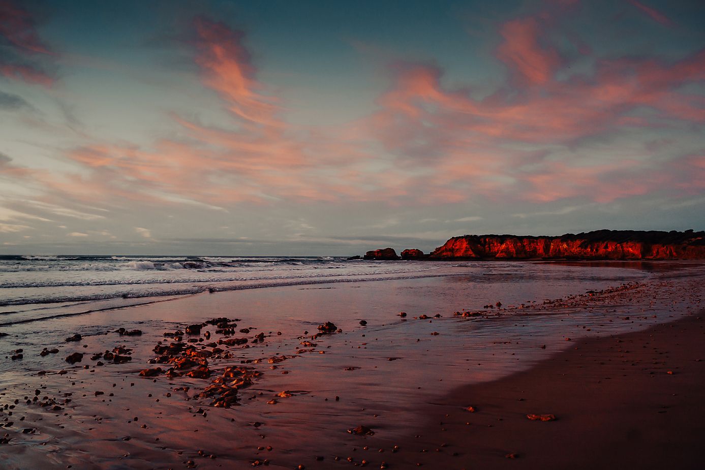 A stunning pink and orange sunset illuminates the coastline as waves crash against the rocky beach.
