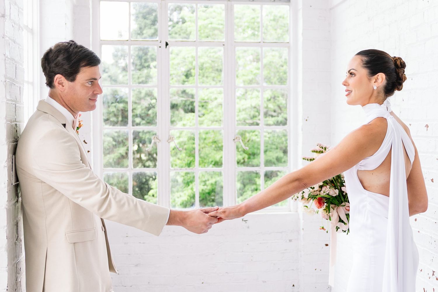 A bride in a white strapless gown poses gracefully by an ornate fireplace in a room with floral wallpaper.