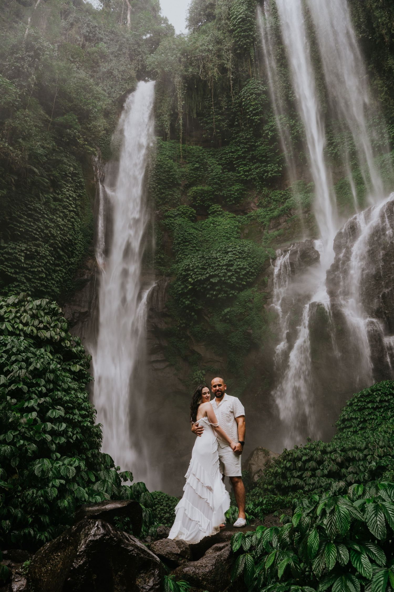 Majestic twin waterfalls cascade down a rocky cliff surrounded by lush tropical plants in a misty forest setting.