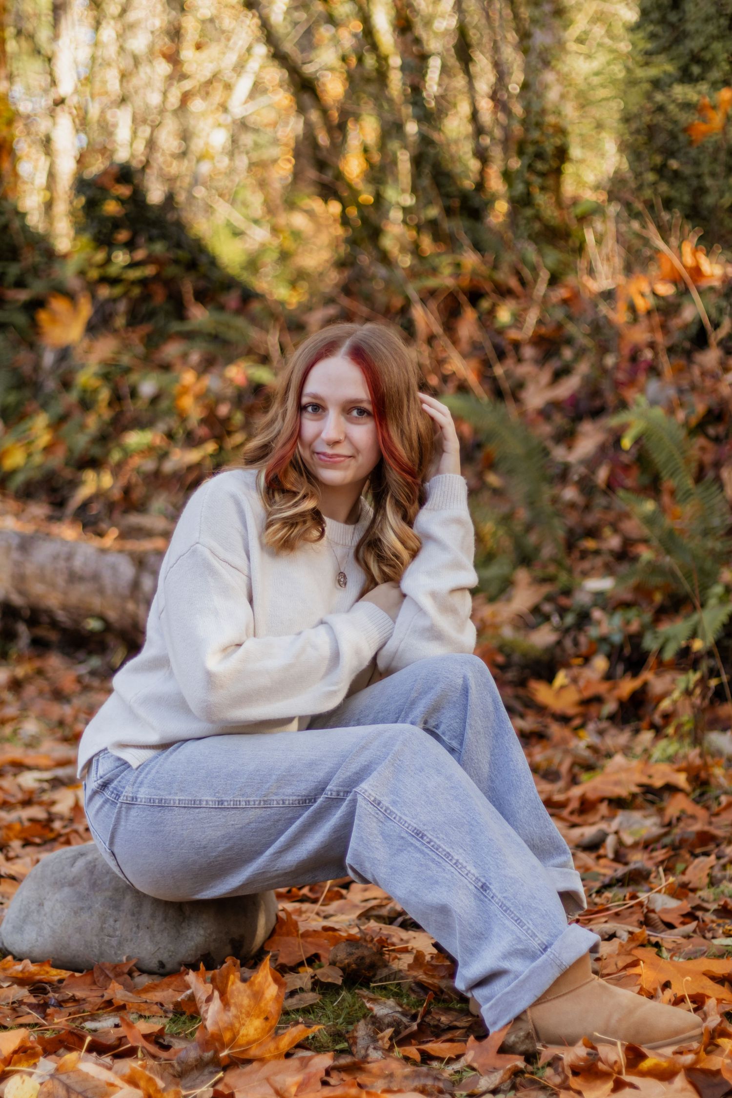 Young woman in cream sweater and jeans sitting on rock in autumn forest.
