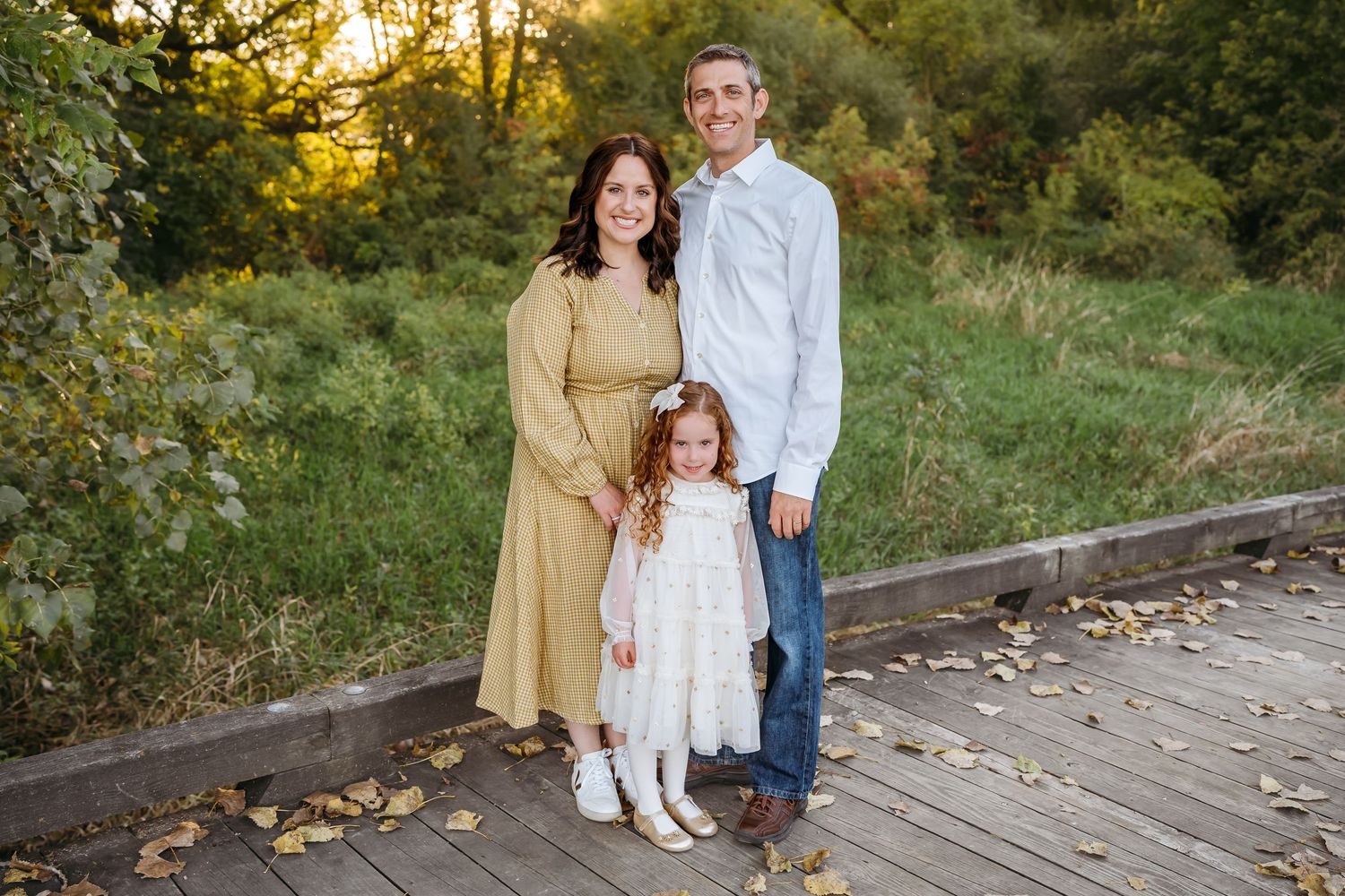 A family of three poses outdoors on a wooden bridge during golden hour with autumn leaves and greenery in background.