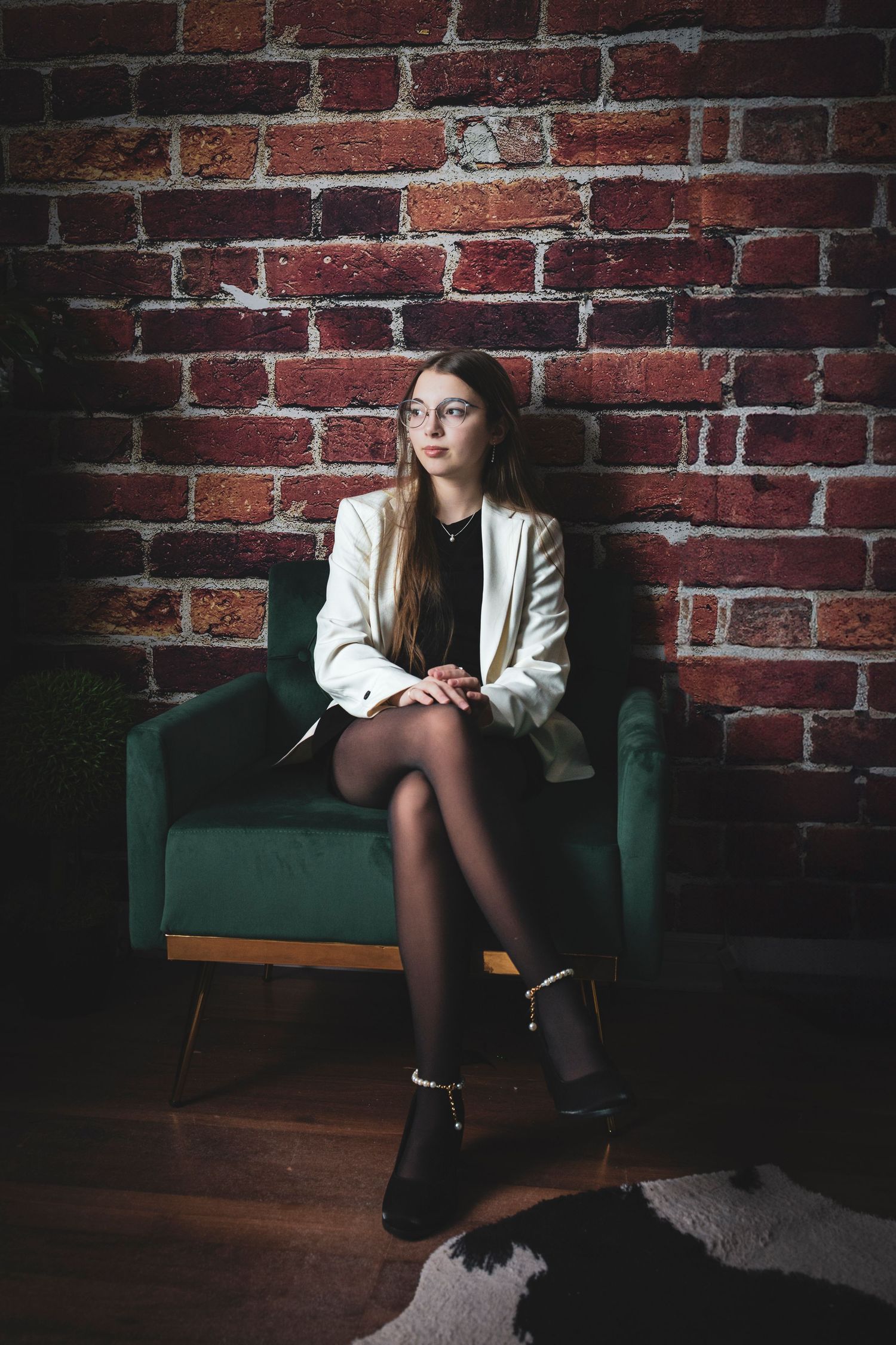 In moody lighting, someone wearing white sits on a teal velvet couch against an exposed brick wall backdrop.
