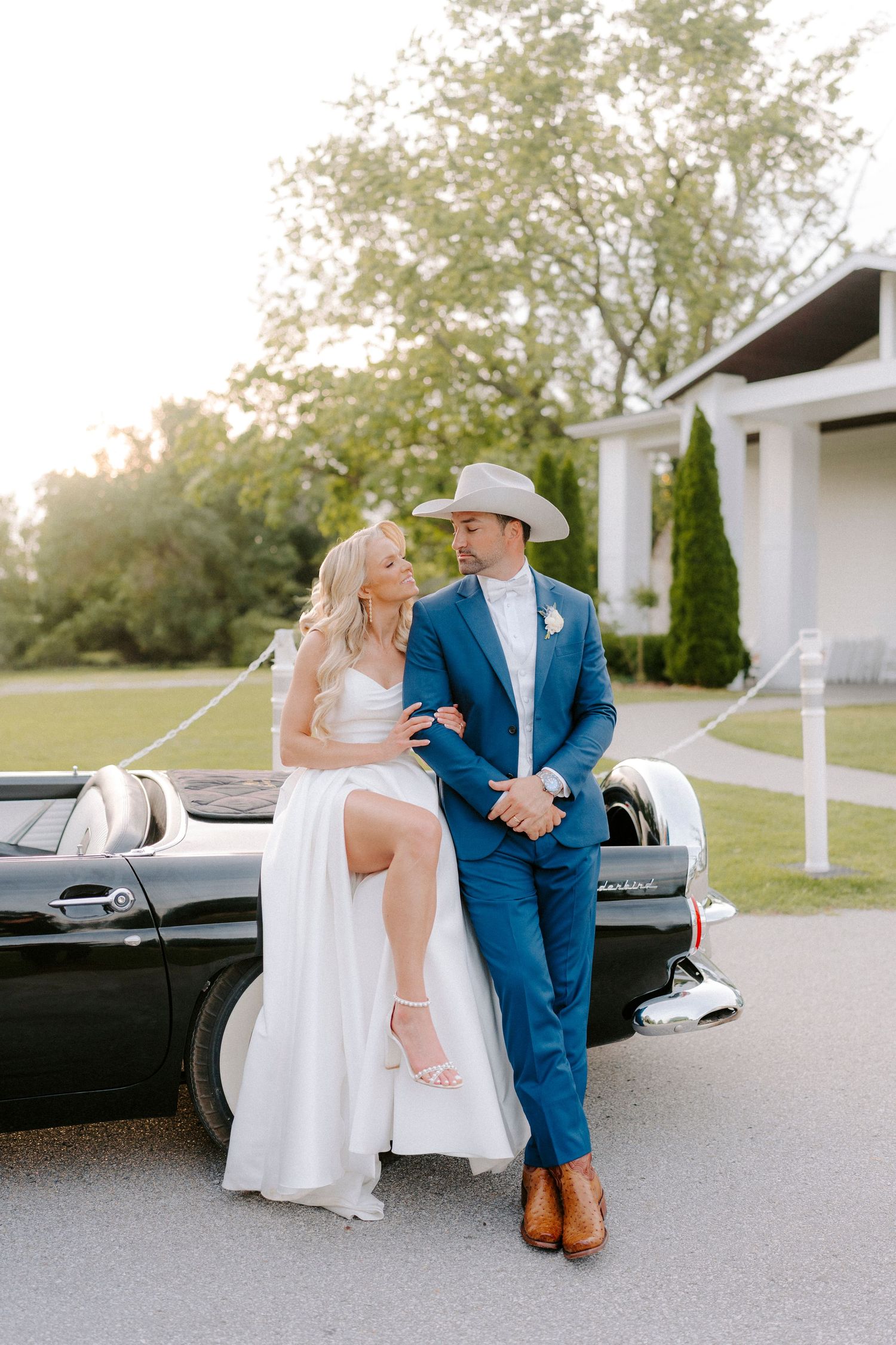 A couple in wedding attire share a romantic moment on a driveway, with the groom wearing a blue suit and cowboy hat.