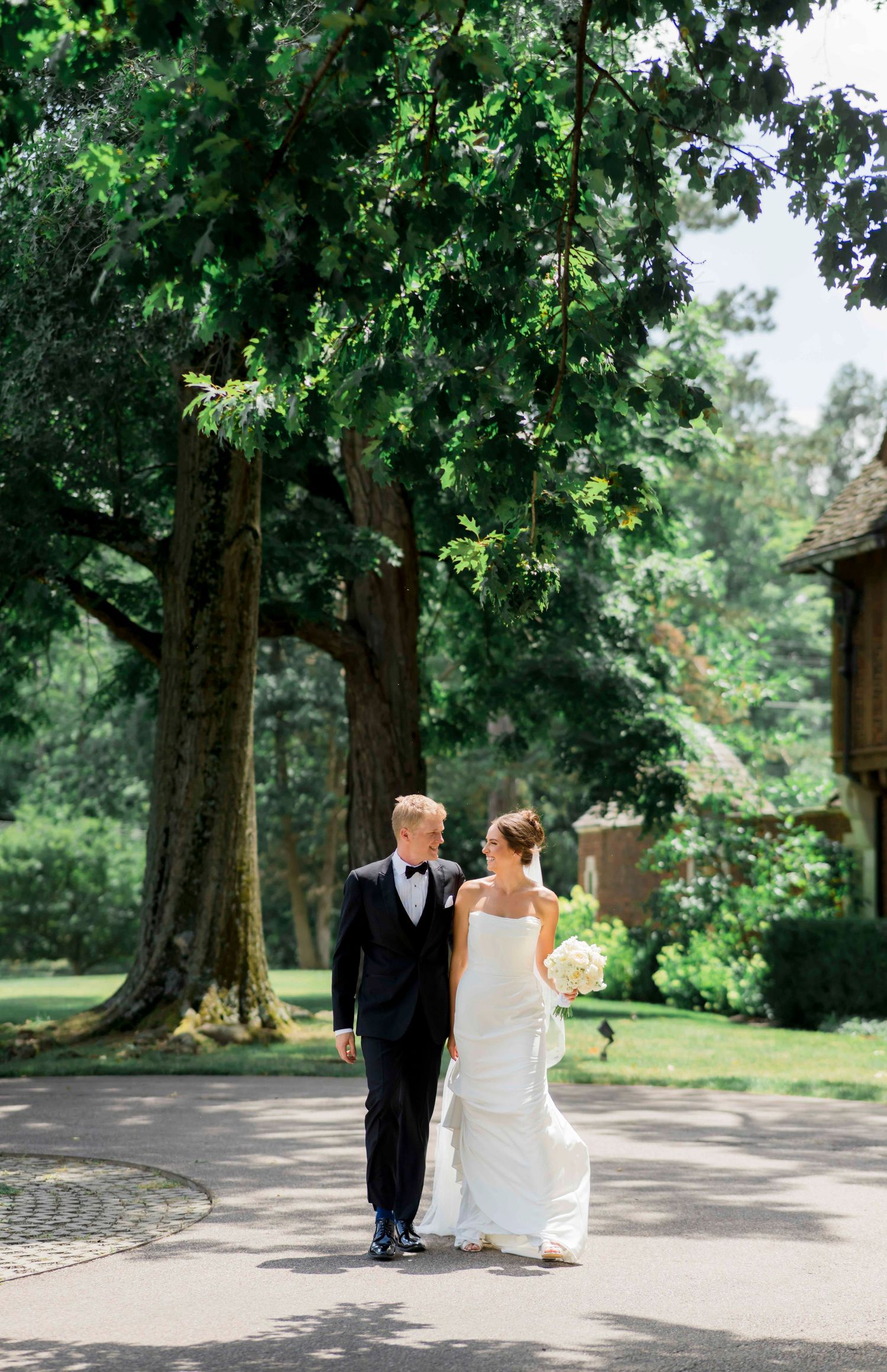 A couple in wedding attire walks along a tree-lined path in a lush garden setting on their special day.