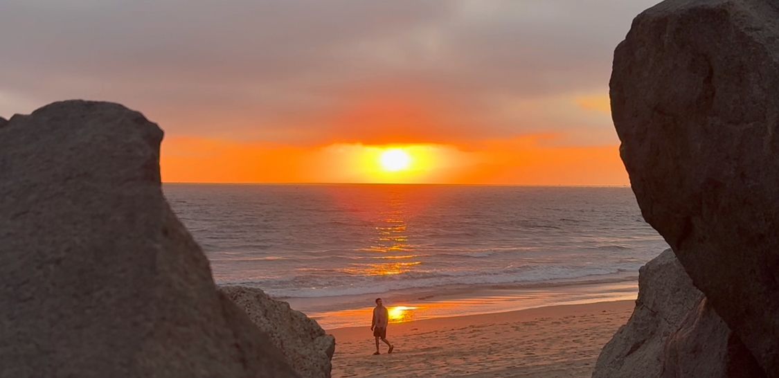 A silhouette stands between rock formations overlooking a vibrant orange sunset at the beach.