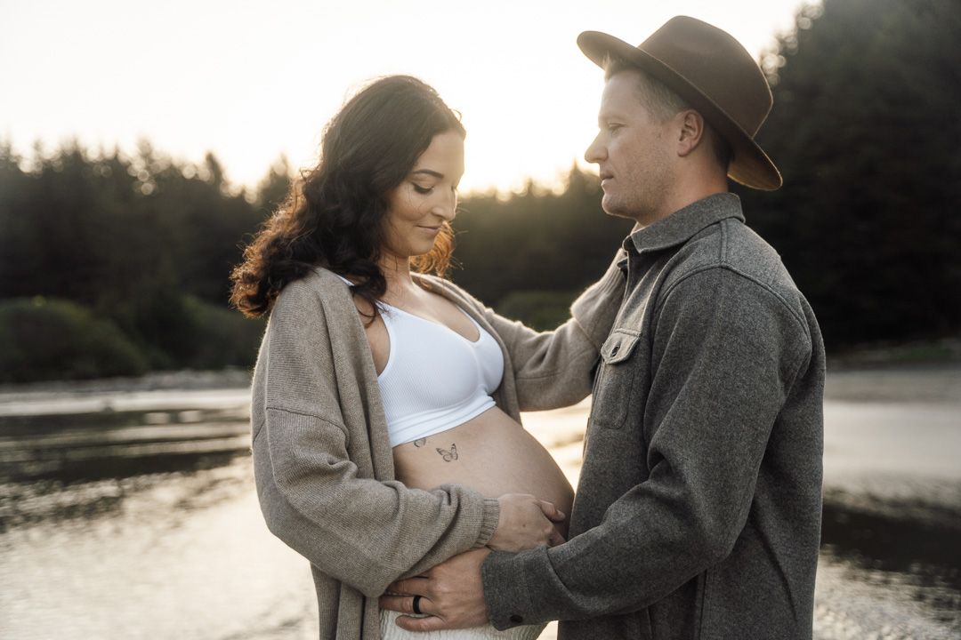 A romantic maternity photo of a couple embracing by a lake at sunset wearing cozy sweaters in Arlington Heights