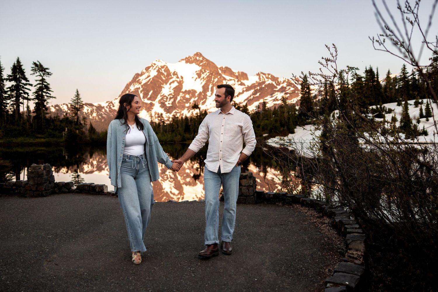Couple walks together on mountain trail with snow-capped peaks glowing orange at sunset in the background.