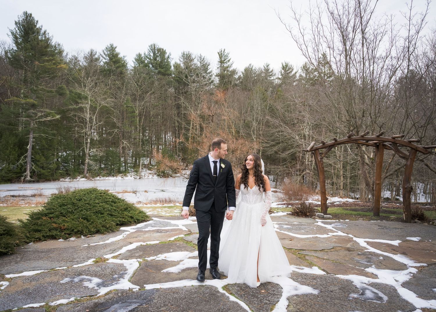 A couple in wedding attire stands in a snowy landscape surrounded by evergreen trees and melting patches of snow.