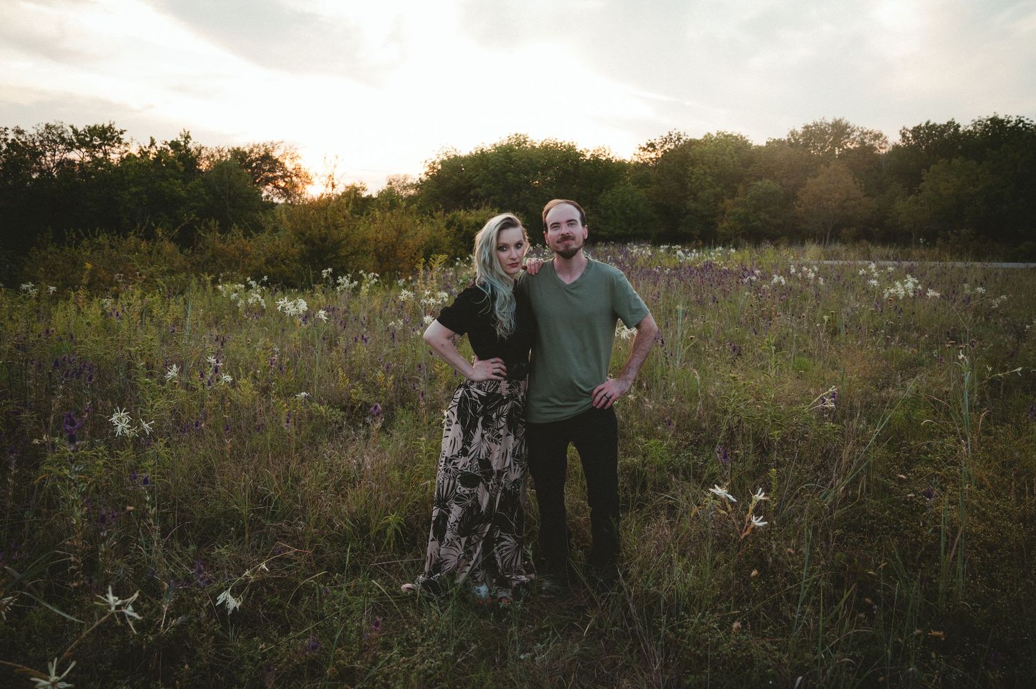 A couple (Jon and Heather photography team) stands together in a golden meadow at sunset surrounded by wildflowers and tall grass.