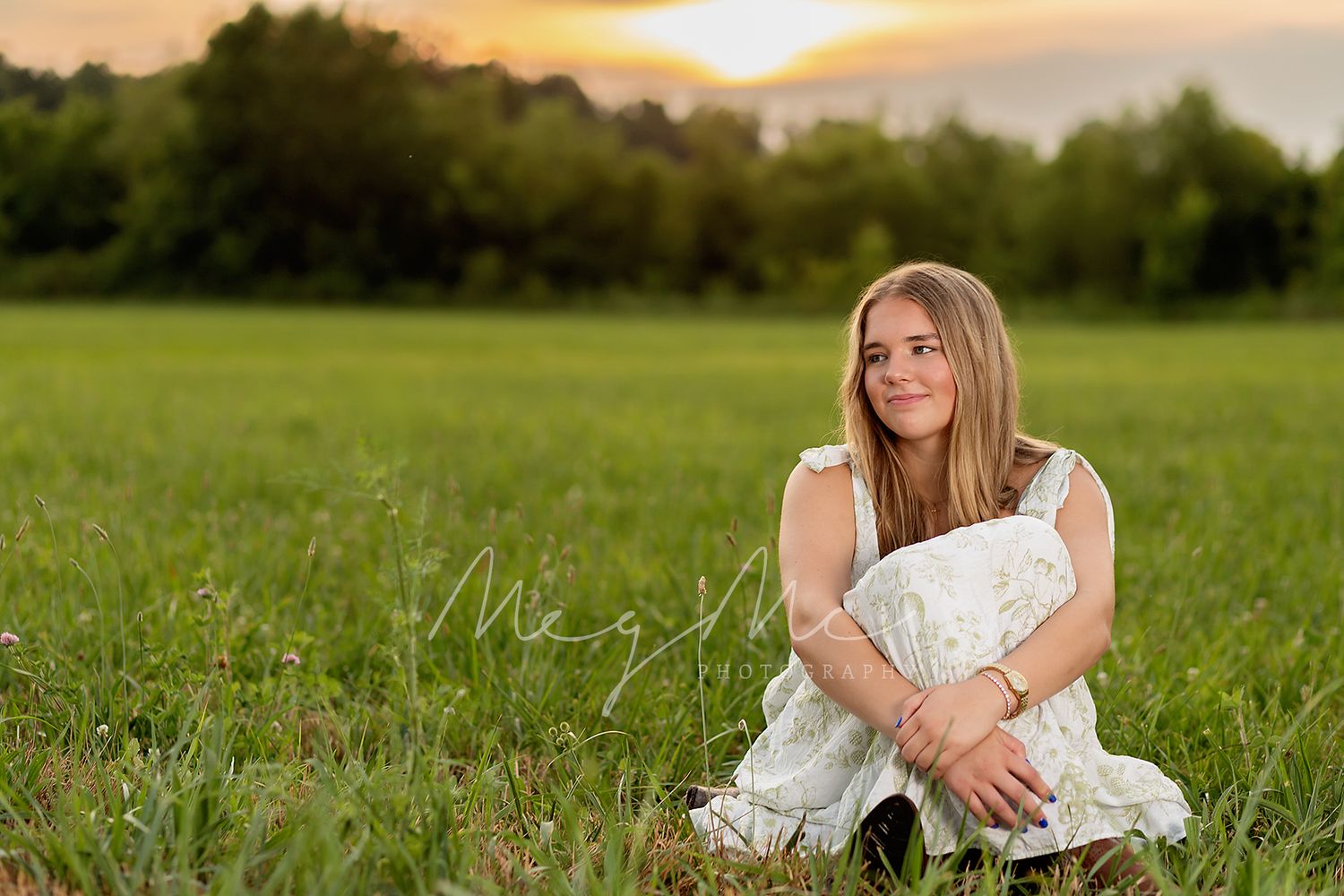 A female teen in a white summer dress sits thoughtfully in a green meadow at sunset. Senior Pictures with MegMc Photography