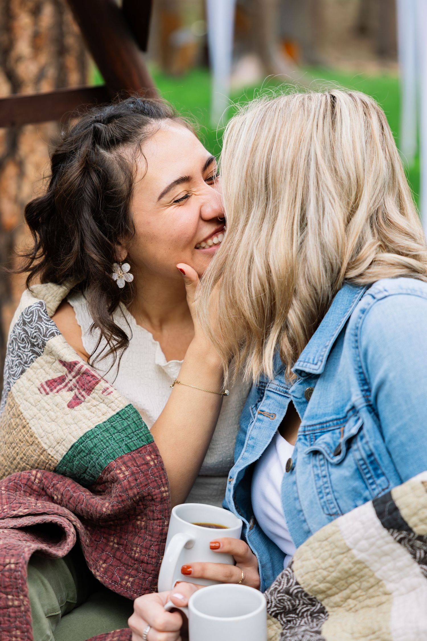 Two women sharing an intimate moment outdoors while holding coffee mugs and wearing casual outfits with denim and knits.