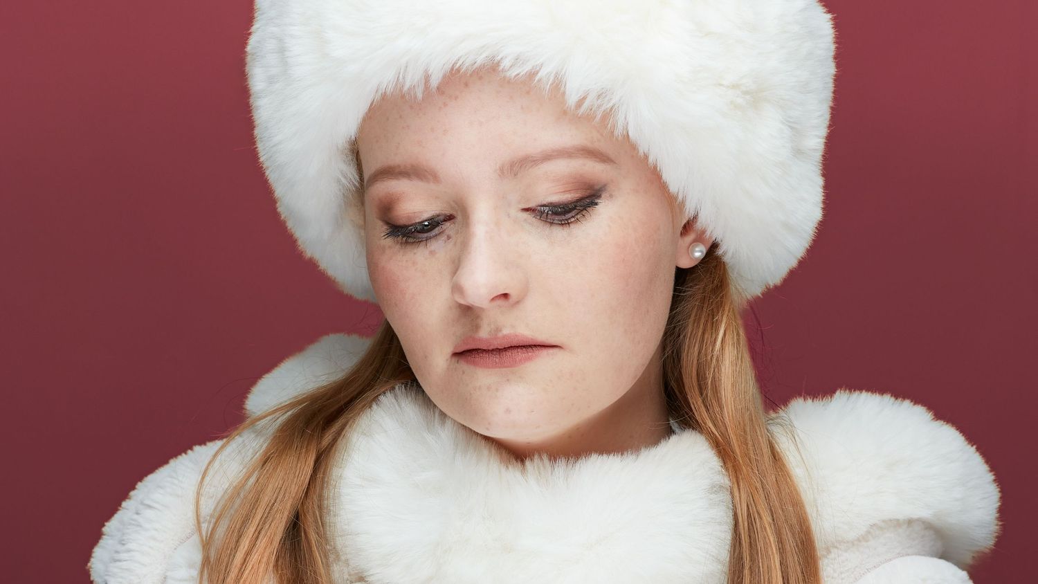 A person wearing a fluffy white winter hat poses for a series of expressive photos against a burgundy background.