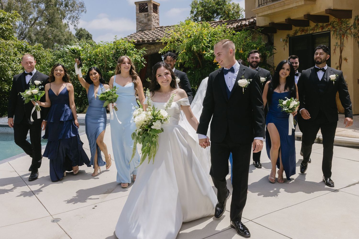 Elegant outdoor wedding procession walking along covered stone path with greenery and flowers overhead.