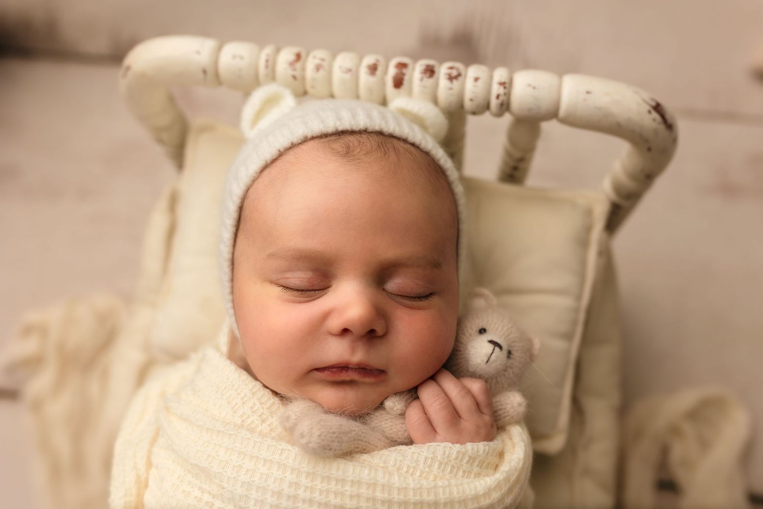 Newborn baby boy sleeping in a spindle bed, wearing a white bear ear hat and holding a teddy bear during a wrapped mini newborn session.
