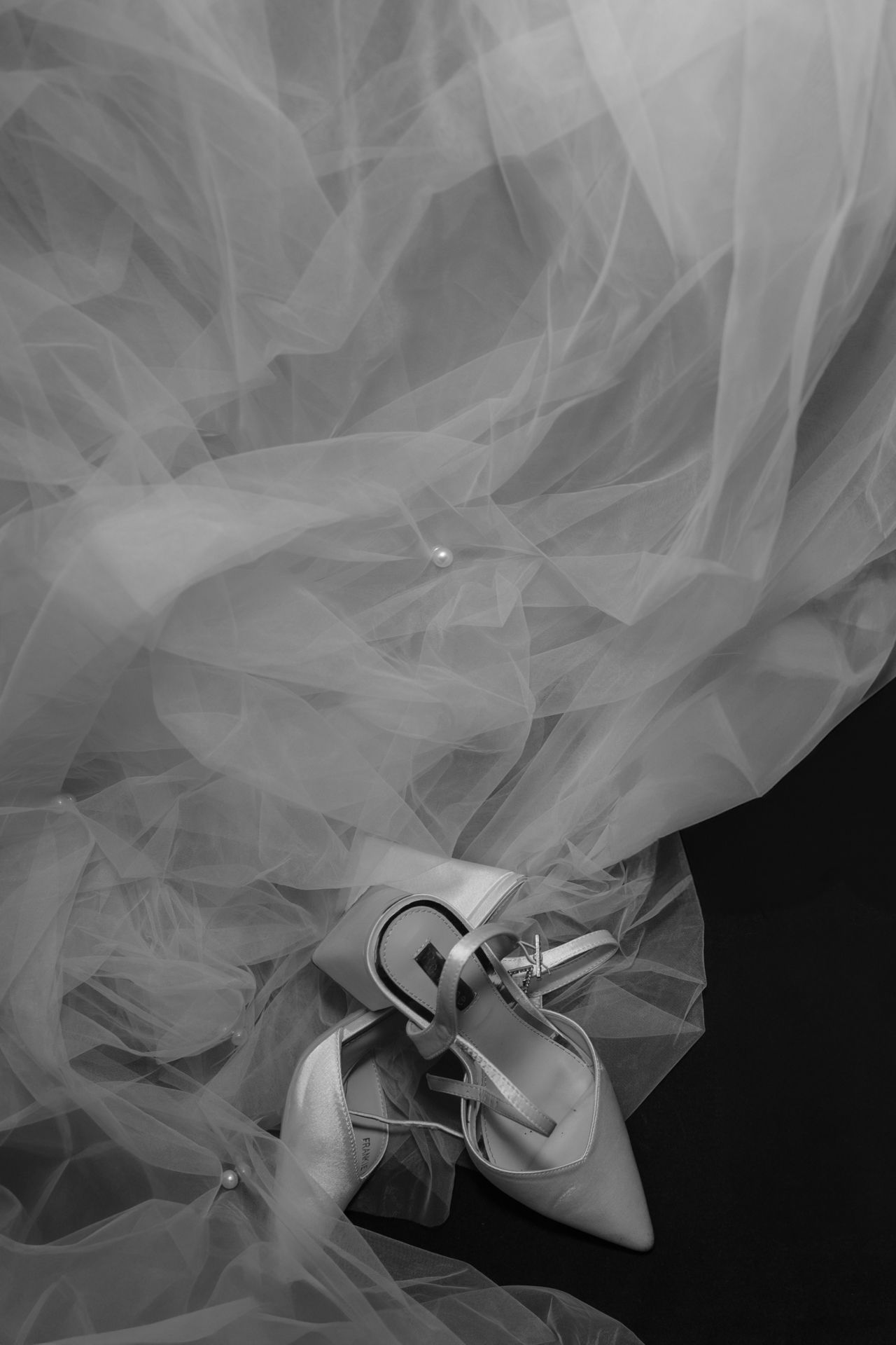 A pair of elegant white satin wedding shoes nestled in layers of flowing tulle in a black and white photograph.