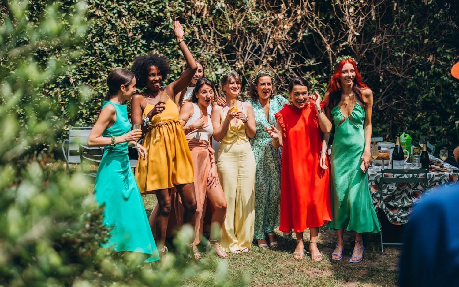 A group of wedding guests in colorful dresses and attire pose together for photos in an outdoor garden setting.