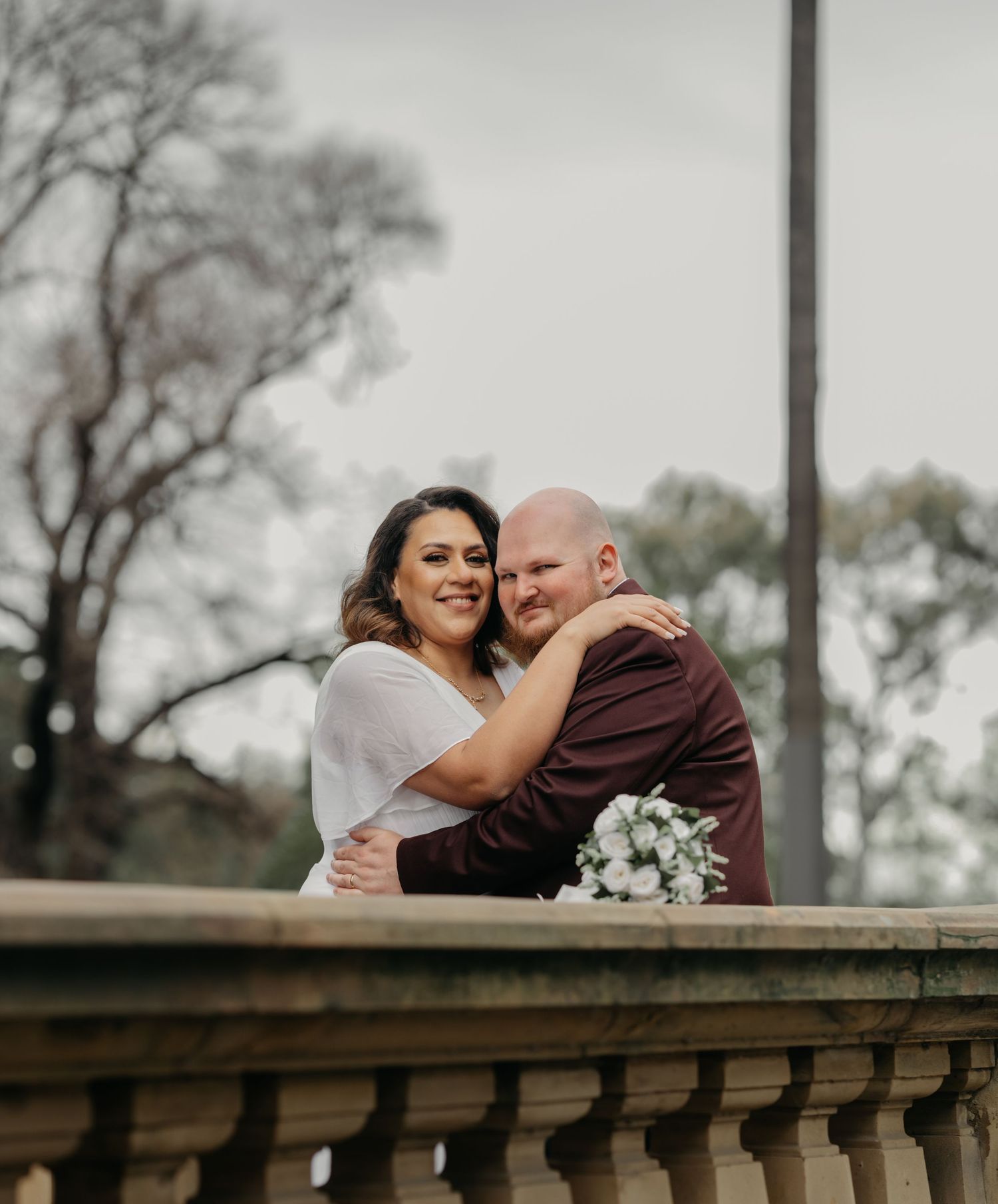 A couple shares a tender embrace while standing near a stone balustrade in an outdoor setting.