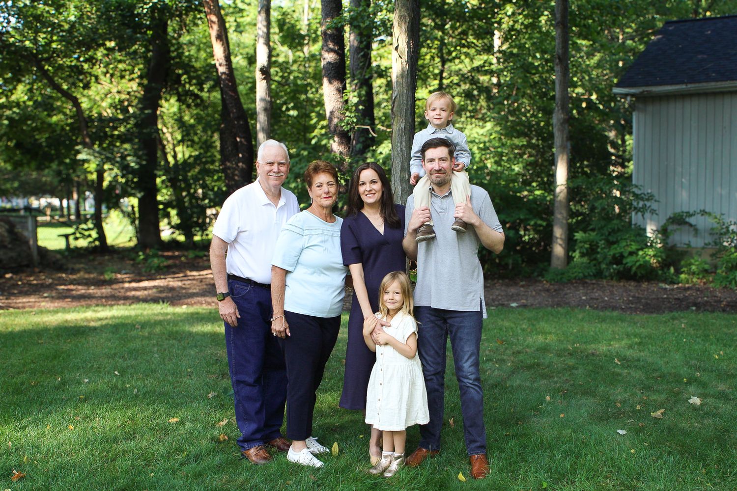 Extended family gathers for outdoor portrait session in park setting with green trees.