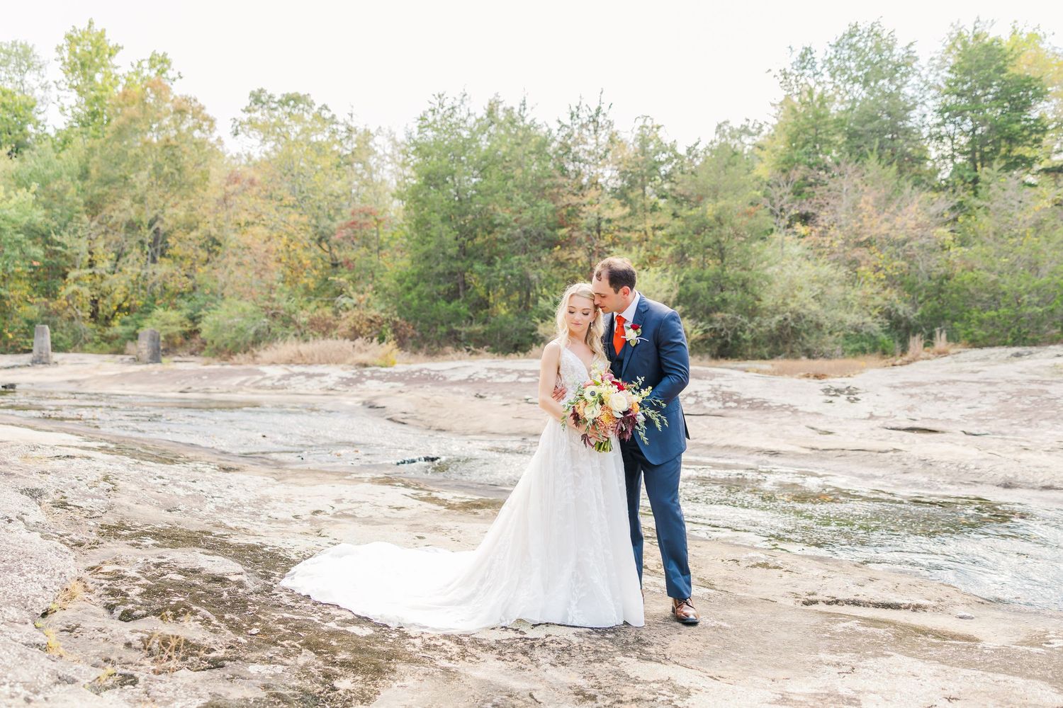 A couple in wedding attire embrace on a rocky terrain surrounded by autumn trees in the background.