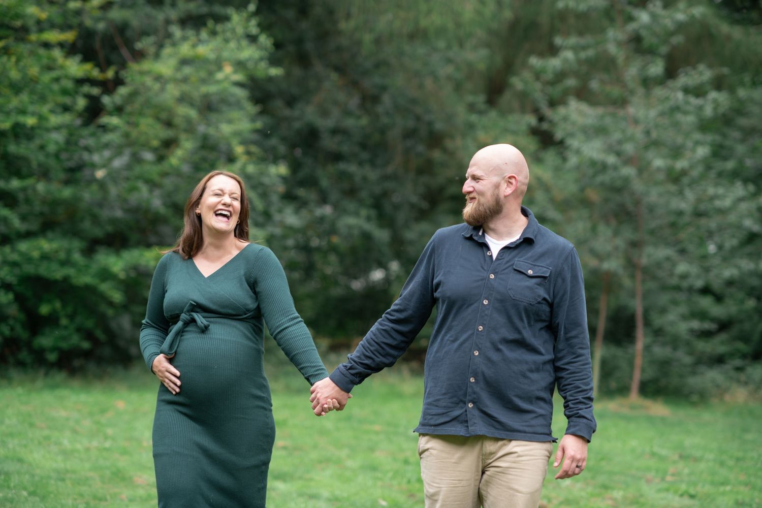A couple walks hand in hand through a lush green park setting during an outdoor maternity photo session.