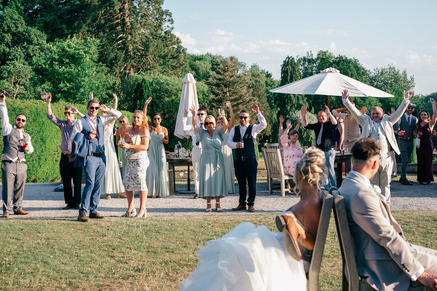Groups of people gather outdoors under umbrellas and pine trees at a sunny outdoor reception or celebration with Nova Scotia photographer