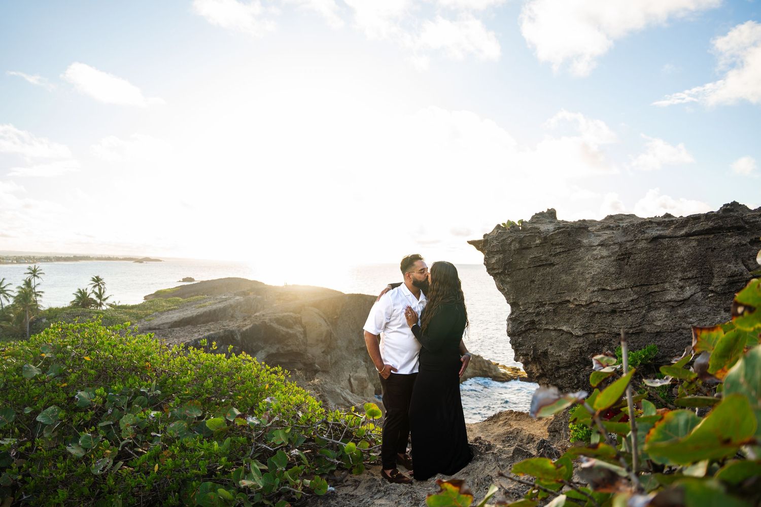 Couple stands on rocky cliff overlooking ocean at sunset with yellow wildflowers in foreground.