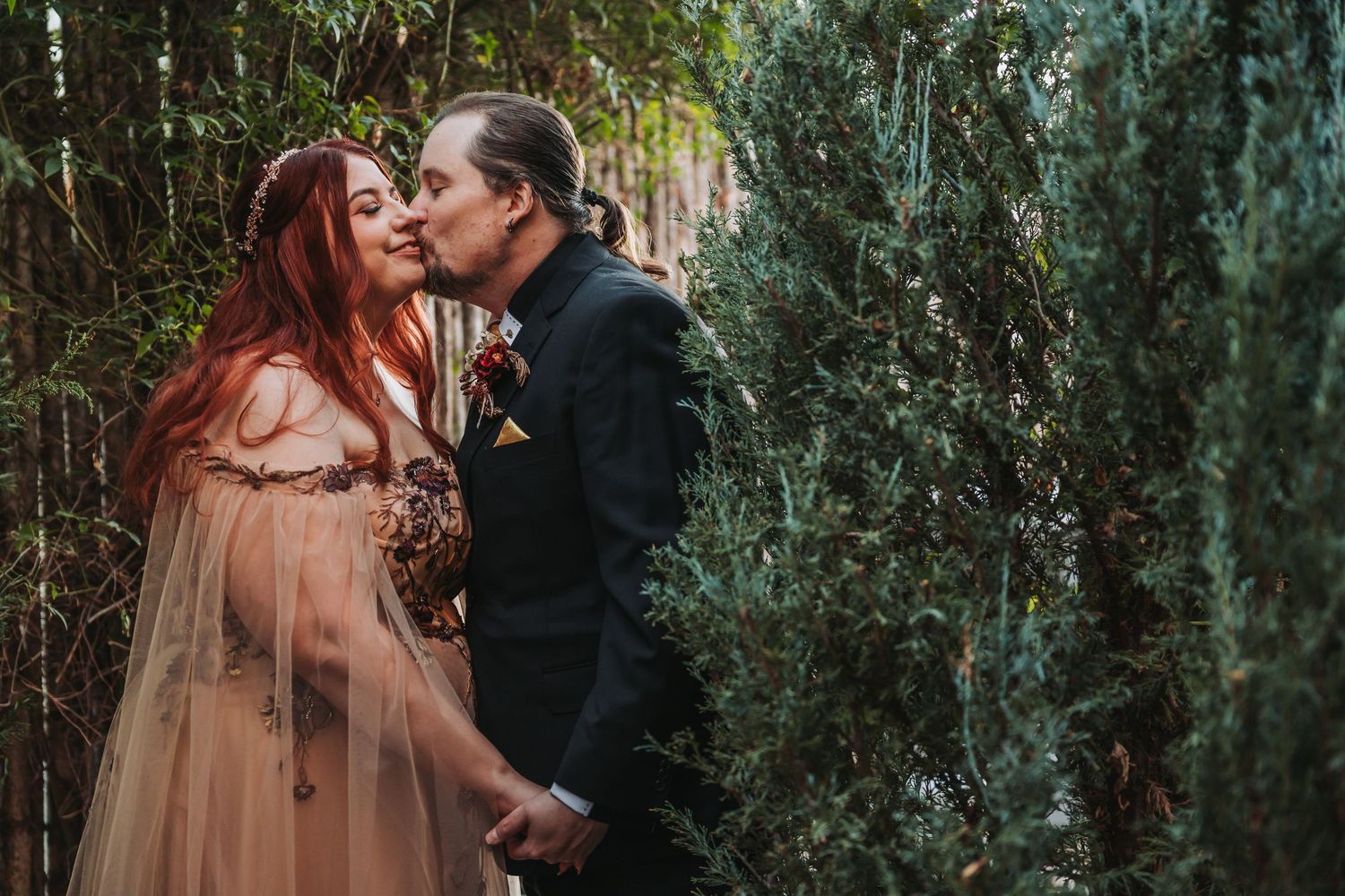 A couple shares a romantic moment in a garden with evergreen trees creating a moody backdrop.