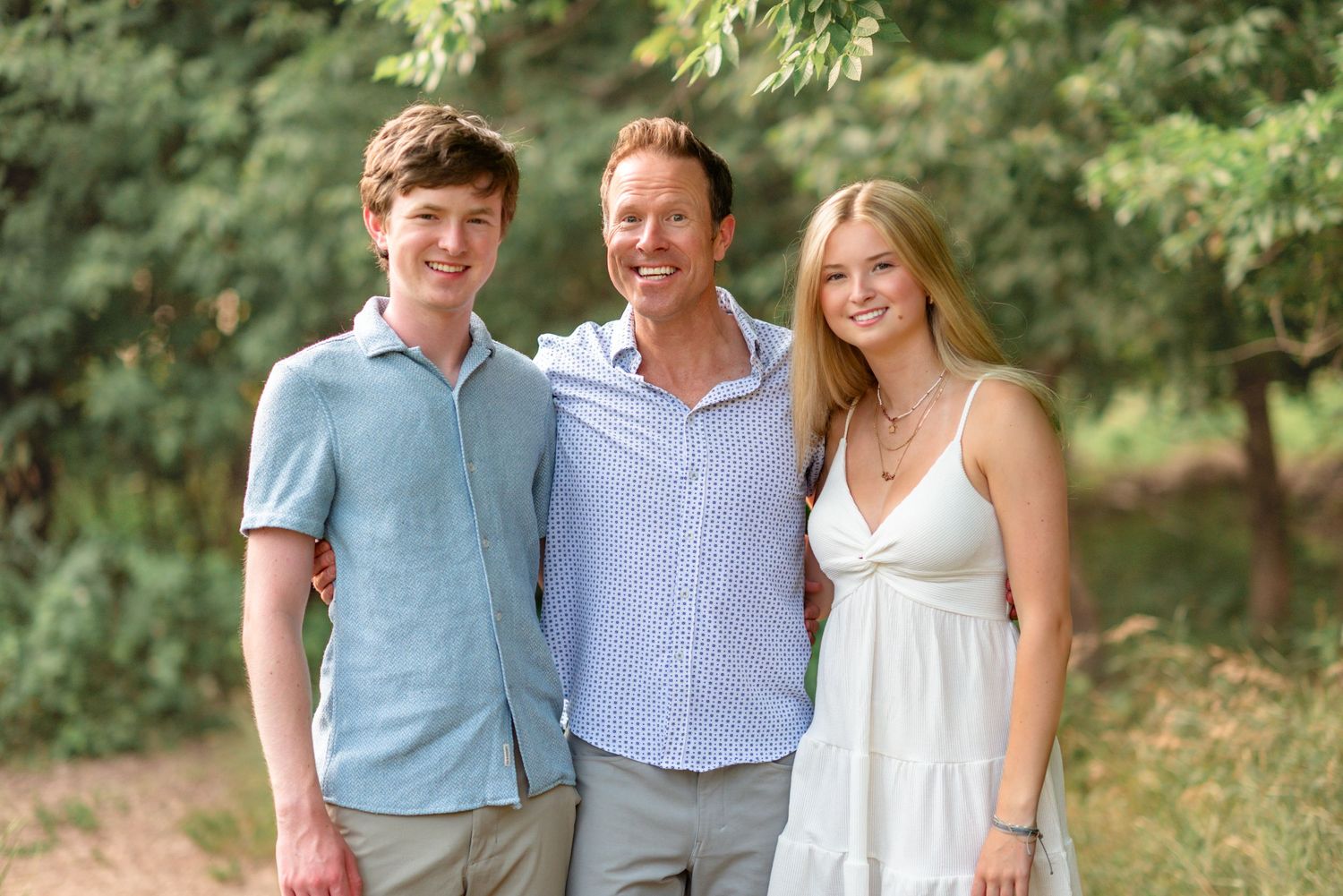 Three people stand together smiling outdoors with green foliage in the background.
