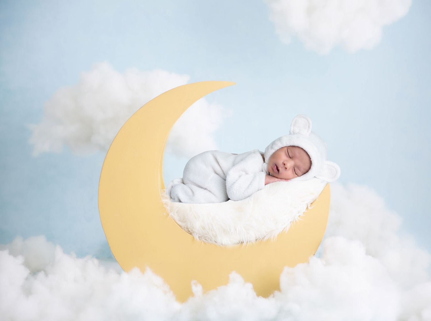Newborn baby sleeping peacefully on a decorative yellow crescent moon against a dreamy cloudy sky background.
