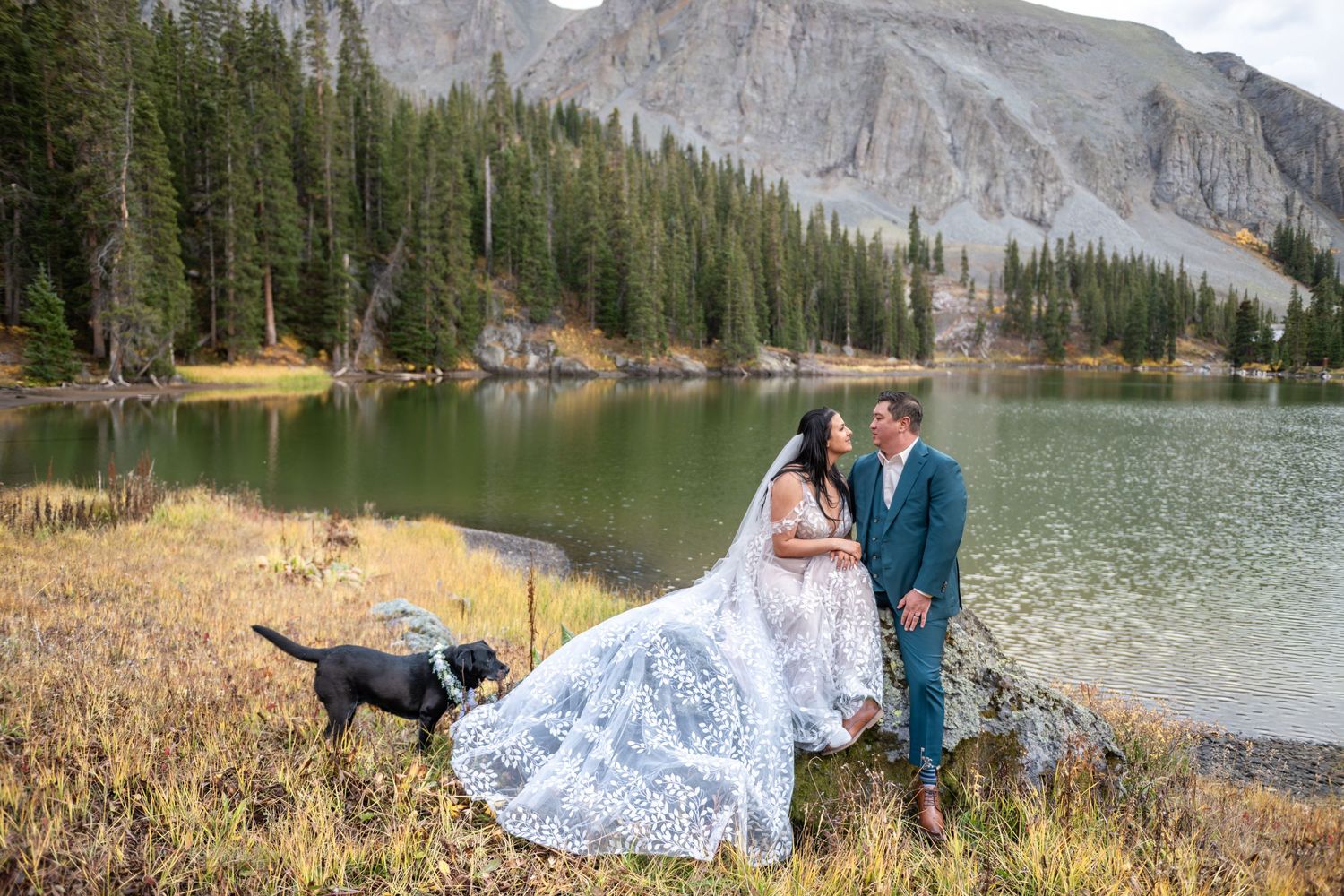 A bride and groom sit side by side on a rock by an alpine lake with their dog by their side. They are looking at one another lovingly.