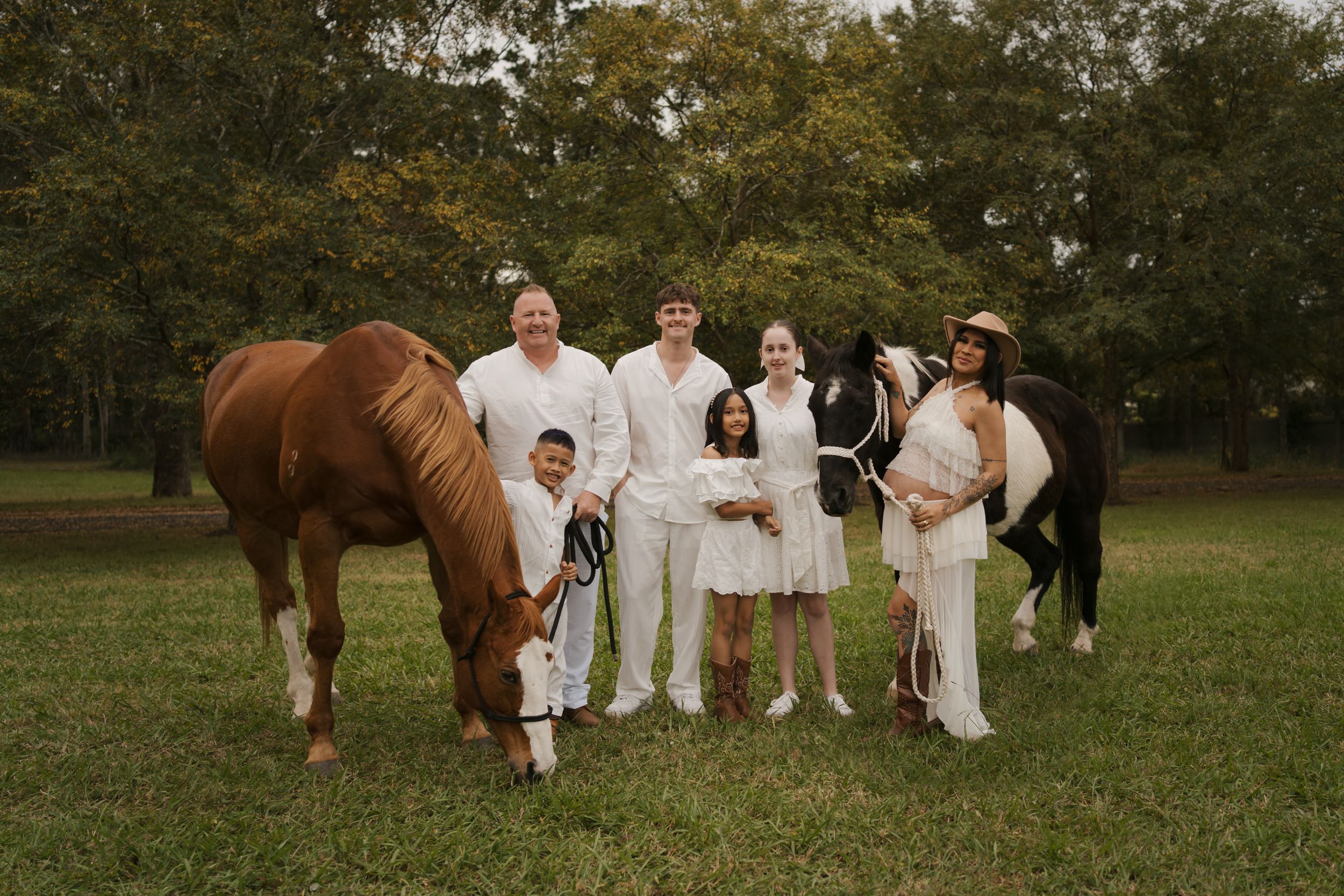 Family in white outfits posing with two horses on a green field surrounded by trees.