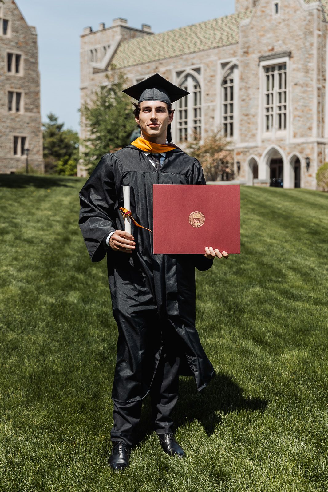 Young man in black graduation gown and cap holds diploma cover on college campus lawn.