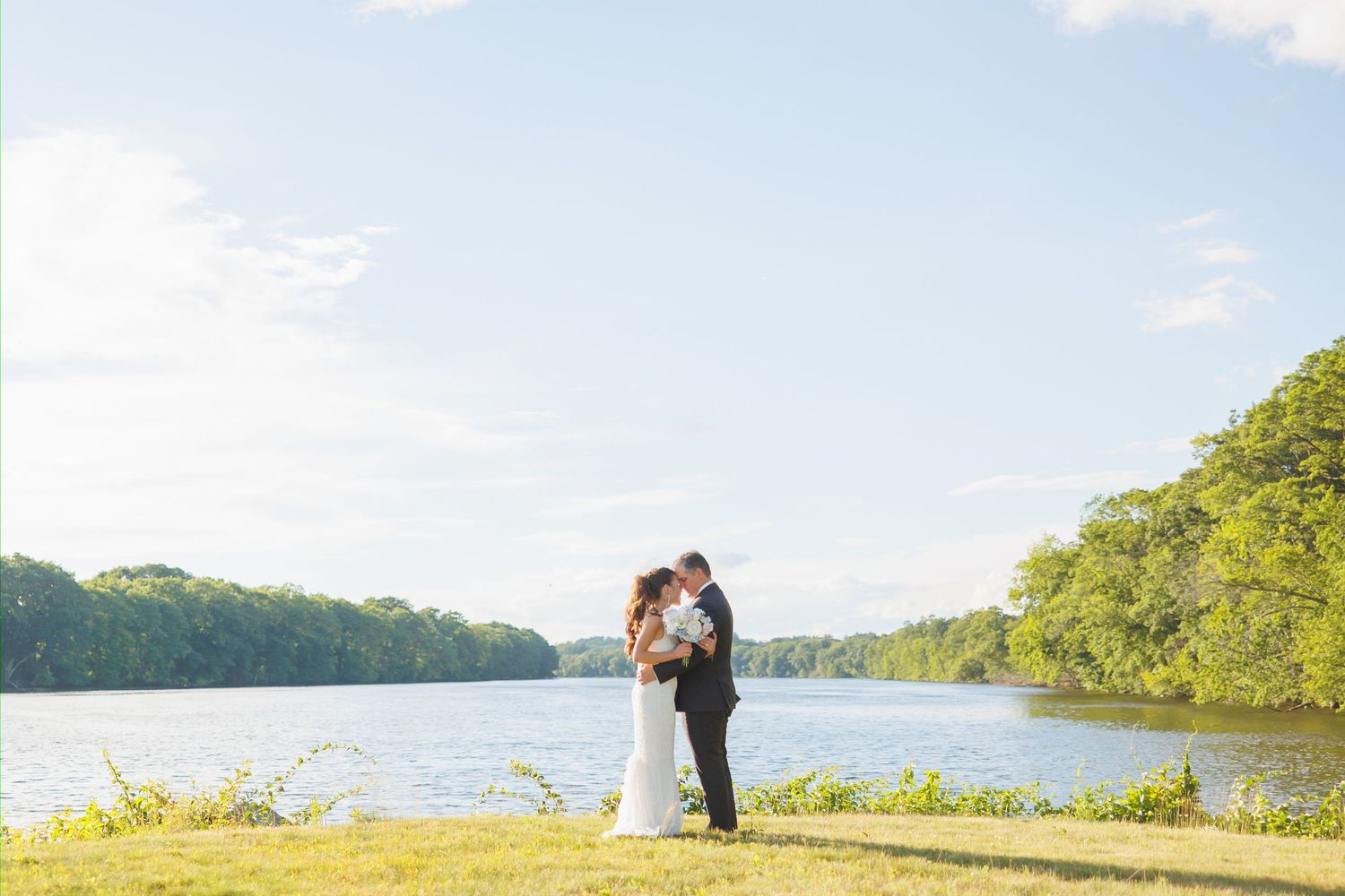 Bride and groom share a romantic embrace by a serene lakeside surrounded by lush green trees on their wedding day.