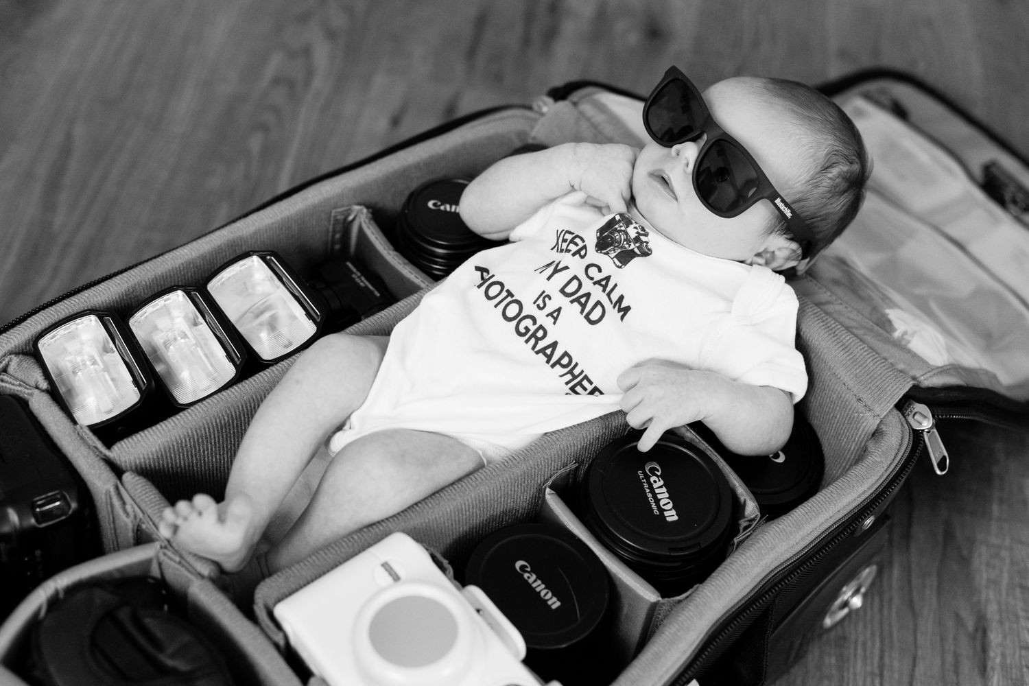 Baby in sunglasses lying in a suitcase surrounded by Canon camera lenses, wearing a photographer dad onesie.