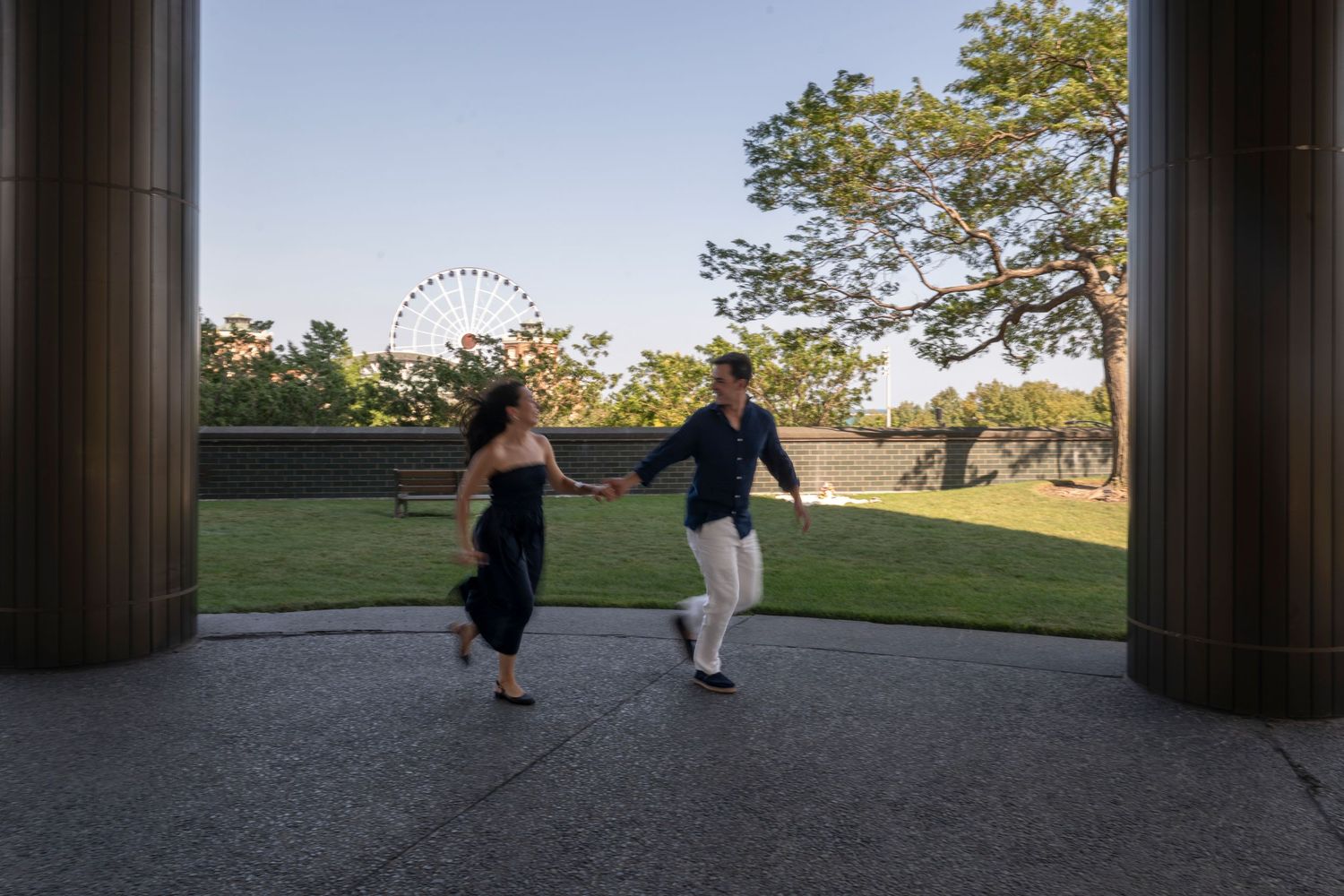 A young couple run across a patio in Chicago with Navy Pier's Centennial Wheel and trees in the background under a blue sky.