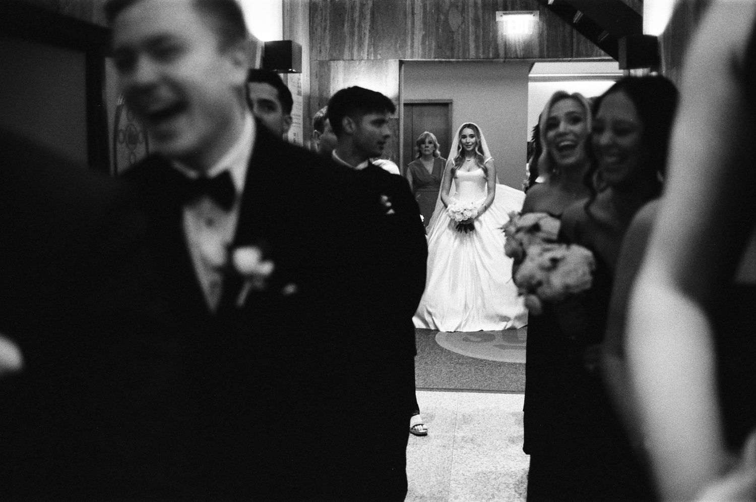 Black and white wedding photo of bride in white gown with guests and groom in tuxedo in foreground.